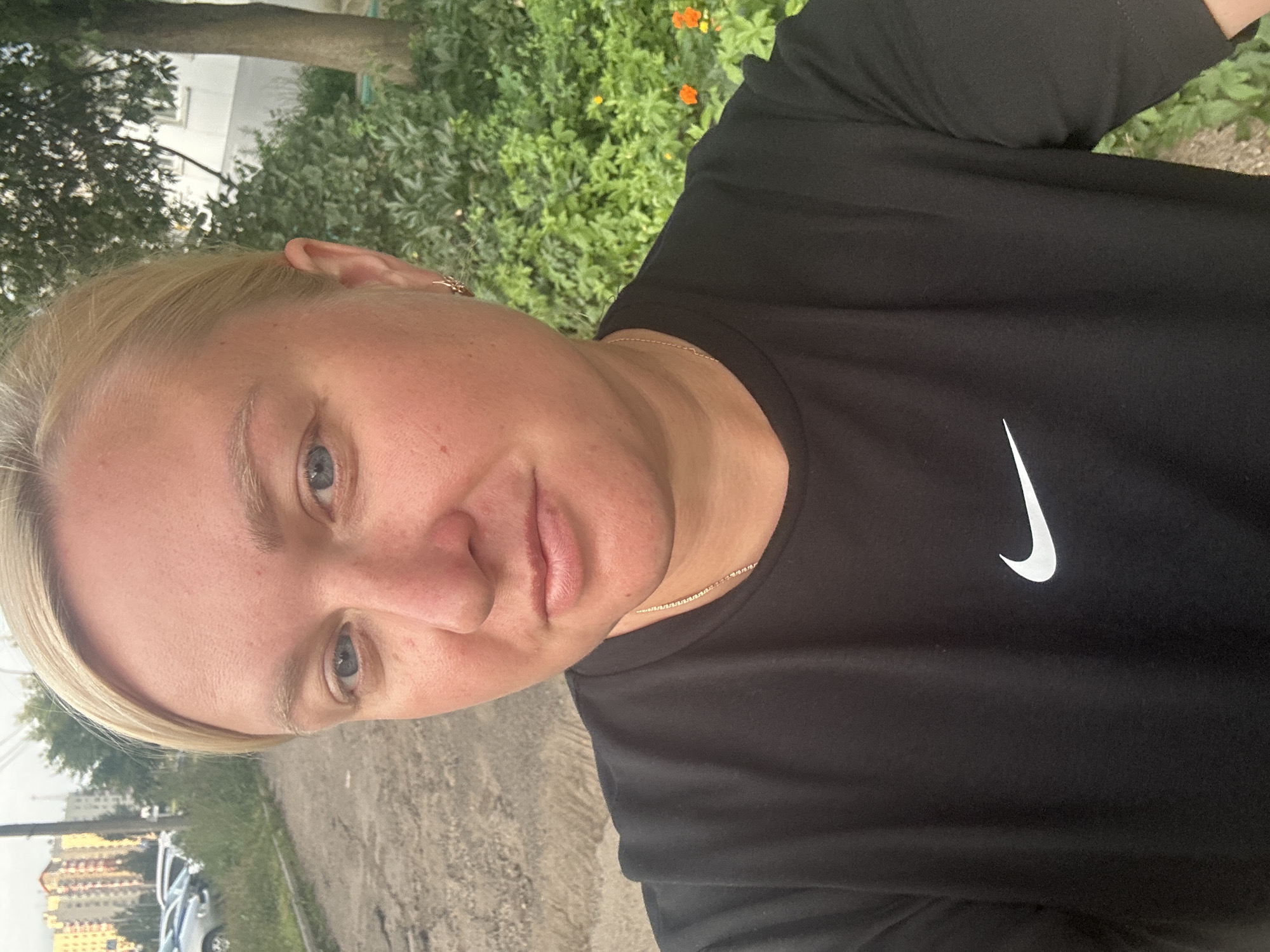 A woman with blonde hair is taking a selfie outdoors. She is wearing a black t-shirt with a white Nike logo on the left side of her chest and a thin gold chain. She has light blue eyes and fair skin. The background shows a paved area, some green bushes with orange flowers, a tree, and buildings in the distance. The overall setting appears to be a residential area in Chaikovsky, Russia, possibly a courtyard or sidewalk area.