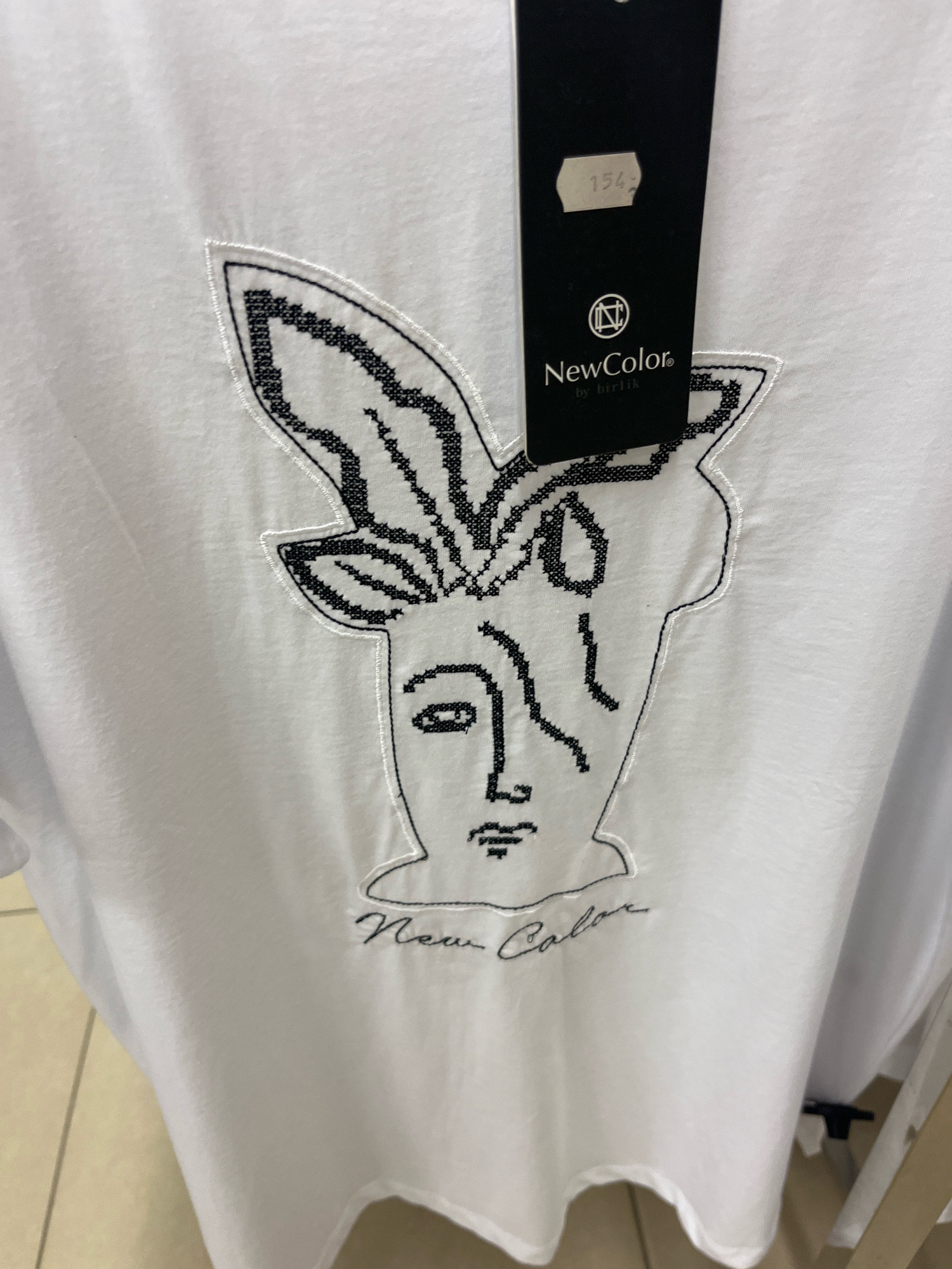 A white t-shirt with an abstract black embroidered design is hanging, possibly in a retail setting. The embroidery depicts a stylized face with what appears to be bunny ears. A black tag is attached to the shirt, displaying the brand name "NewColor" and the text "by birlik." A price of "154" is visible on the tag. The background is a plain, light-colored surface, possibly a wall or clothing rack. The lighting is even and diffuse.