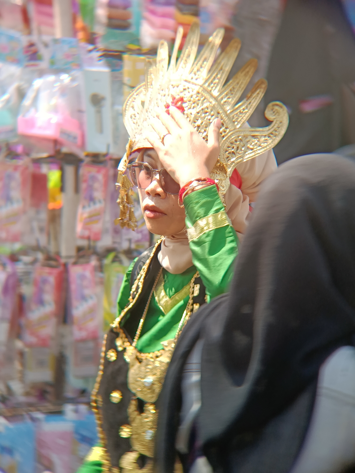 In Desa Kebon Manis, Kecamatan Cilacap Utara, Indonesia, a person is captured from the chest up, adorned in traditional attire. The subject wears a gold headdress with intricate detailing and a prominent, ornate crown that extends upwards. A gold necklace with attached charms is visible around the neck, layered over a vibrant green garment with gold trim on the sleeves. The individual is also wearing sunglasses and a light-colored head covering beneath the headdress. A portion of another person wearing a dark head covering is in the foreground, obscuring part of the main subject. The background is blurred but suggests an outdoor market setting with shelves stocked with various items, possibly merchandise or decorations. The lighting indicates a sunny day.