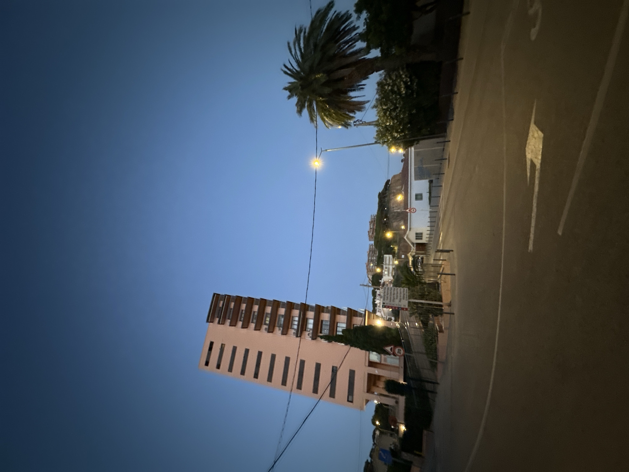 The image captures a twilight scene in Sant Feliu De Guíxols, Spain. A tall, slender, peach-colored apartment building with horizontal dark-framed windows and balconies dominates the left side of the frame. In the background, a palm tree stands to the right of the apartment building, with other greenery and buildings on a hillside. A streetlamp illuminates the area, casting a warm glow on the surrounding road and pavement, which features white lane markings, including an arrow indicating direction. The sky is a deep blue, transitioning from the darkness of night. Several power lines are visible stretching across the sky. A white rectangular sign with text and a speed limit sign indicating "20" are placed on the side of the road. A single-story white building with red trim is visible on the right side of the frame, near the palm tree. The overall impression is one of a quiet, possibly residential, urban environment at the end of the day.