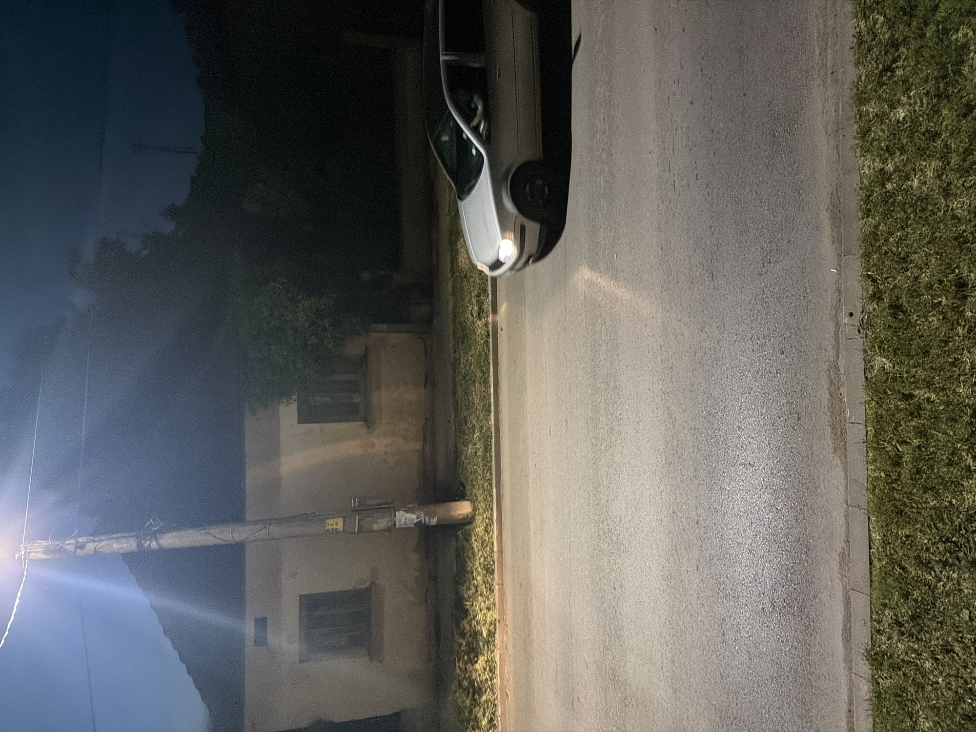 This image depicts a nighttime street scene in Iskar, Bulgaria. The primary subject is a light-colored (possibly white or silver) sedan, seen from the front-left, driving on an asphalt road. Its headlights are on, casting distinct beams of light onto the road surface ahead.
To the left of the road stands a tall utility pole with a bright street light at its top, illuminating the immediate area and creating noticeable lens flares in the photograph. Several utility lines and a large, cylindrical pipe or conduit extend horizontally from this pole towards the background. In the background, partially obscured by dark foliage, are two older, rectangular buildings with visible, unlit windows, suggesting a residential or older commercial setting. Trees and dense greenery form dark silhouettes against the dim, dark blue night sky.
The road itself is paved with asphalt, showing a textured surface. To the right of the road, a strip of green grass, acting as a verge, is visible, separated by a curb. No people are discernible in the scene.
Notable details include the clear night sky, the artificial lighting creating strong contrasts, and the general quiet atmosphere of a street at night. Small, unreadable textual labels or stickers are visible on the utility pole, one appearing yellow, and on the horizontal pipe, which appears to be a torn white label. The image captures a moment of typical evening activity in a suburban or small-town environment.