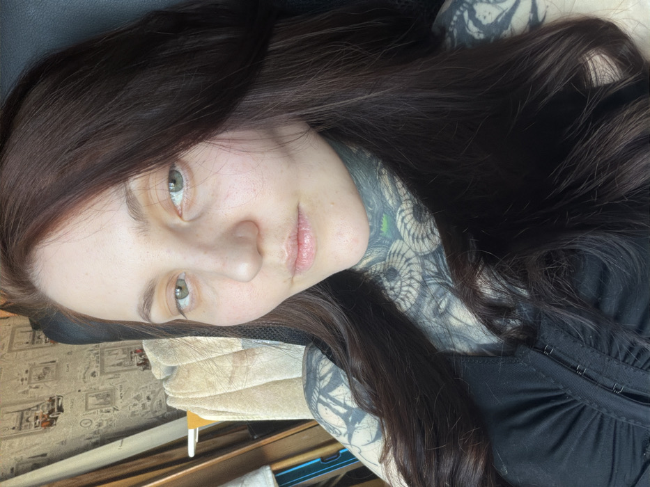 A woman with fair skin, freckles, and light eyes poses with her head tilted. She has long, dark hair. A large, detailed tattoo covers the side of her neck and extends onto her chest and arm. She is wearing a black top with a visible corset-style fastening. The background includes what appears to be a chair and a glimpse of a patterned surface, possibly a desk or floor.