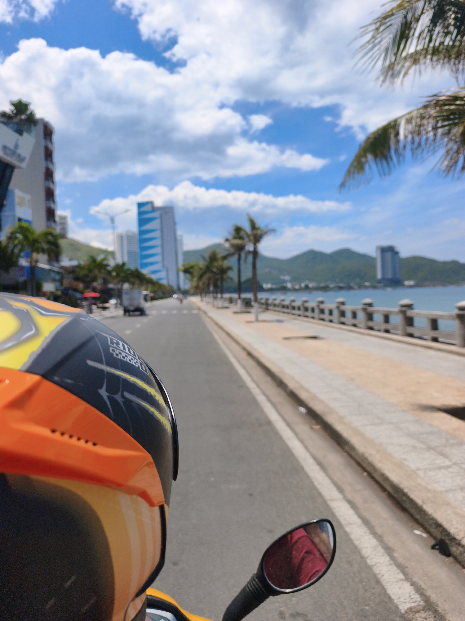 The image depicts a point-of-view shot from the perspective of someone riding a motorbike in Nha Trang, Vietnam. The foreground is dominated by the rider's orange and black helmet. The road ahead stretches into the distance, lined with palm trees and modern buildings, suggesting a coastal urban setting. The sea is visible to the right, with a pedestrian walkway and a decorative railing separating it from the road. In the background, mountains form a scenic backdrop. The sky is blue with scattered white clouds, indicating fair weather. A truck and other vehicles are seen on the road, and a few figures are visible on the sidewalk. The reflection in the motorbike's side mirror shows a person wearing a red garment.