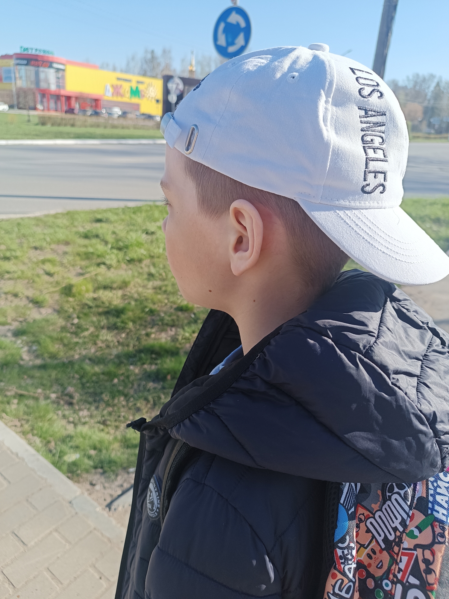 A young boy stands on a sidewalk in Chaikovsky, Russia, facing left. He is wearing a white baseball cap with "LOS ANGELES" embroidered vertically on the side, a black puffy jacket, and a backpack with colorful cartoon stickers. The sidewalk is made of light-colored pavers. Behind him is a grassy area, a road with a roundabout sign, and a large, brightly colored building.