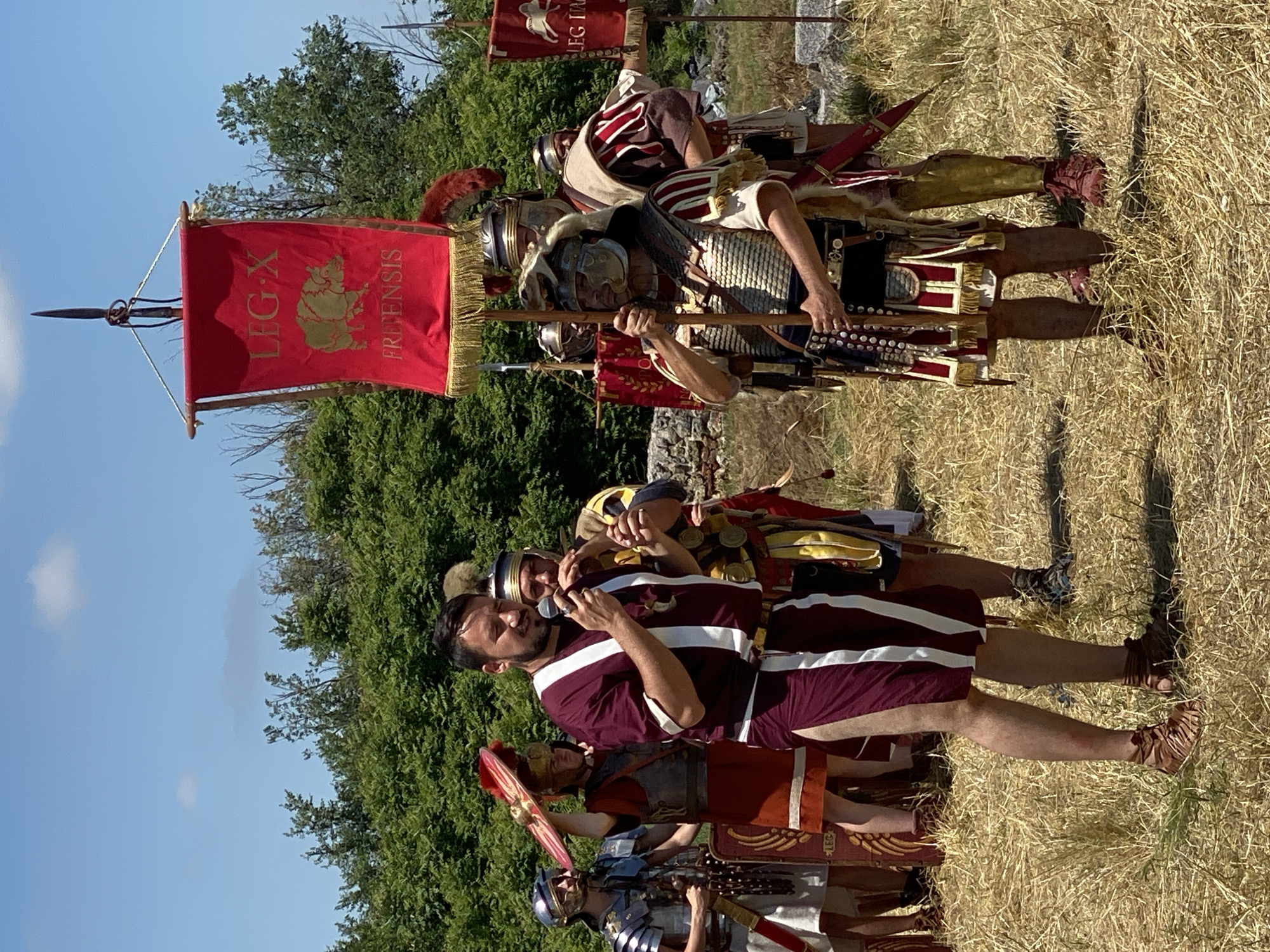 This image, taken in Gigen, Bulgaria, presents an outdoor scene depicting a Roman legionary reenactment under a bright blue sky with scattered clouds. The ground is covered with dry, yellowed grass. In the background, dense green foliage forms a natural wall, with stone ruins partially visible on the mid-right.
At the center of the frame, a man with dark hair and a beard, wearing a maroon tunic with a vertical white stripe and matching shorts, is speaking into a silver microphone. He is oriented slightly to his right. To his immediate right and slightly behind him, another individual in Roman attire is playing a brass wind instrument.
A central figure to the right of the speaker stands prominently, serving as a standard bearer. This individual wears detailed Roman armor, including a helmet adorned with an animal pelt, and holds a tall wooden pole. Suspended from this pole is a rectangular red vexillum with gold fringe, featuring the inscription "LEG X" at the top and "FRETENSIS" at the bottom, flanking a golden wild boar emblem.
Additional reenactors in various forms of Roman armor and tunics are positioned throughout the scene. On the far left, a person in lorica segmentata is partially visible. On the far right, another red banner with the partial inscription "LEG IA" can be seen. The individuals are standing in various poses, suggesting a presentation or performance.