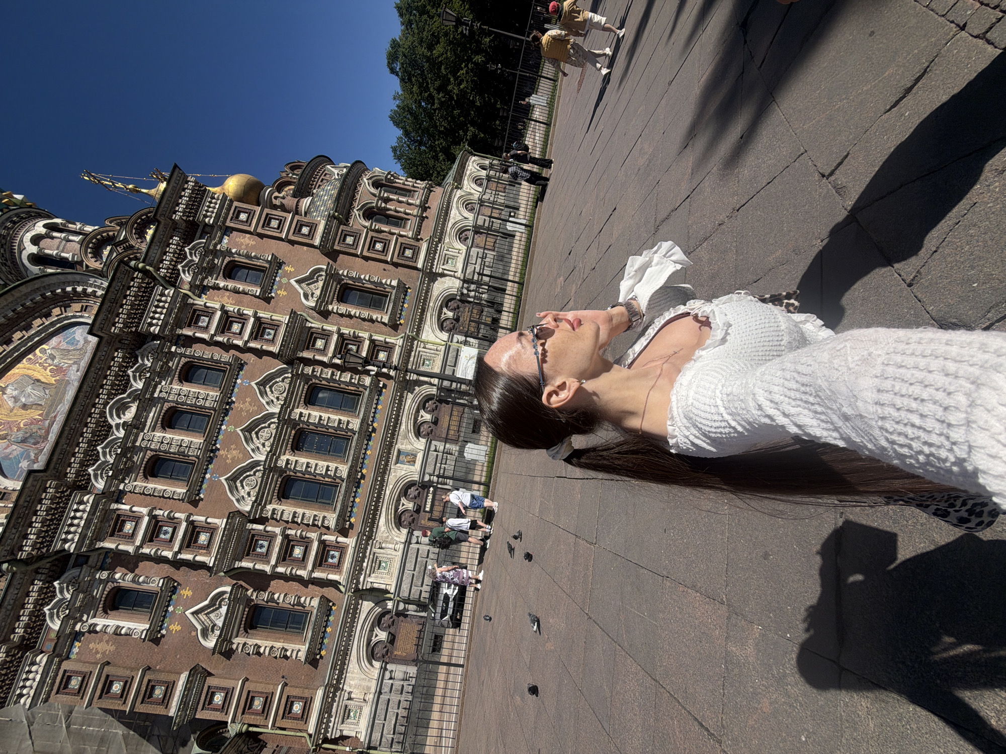 An outdoor wide-angle shot on a bright, sunny day captures a woman in the foreground taking a selfie in front of the Church of the Savior on Spilled Blood in Saint Petersburg, Russia.
The woman occupies the lower-left to mid-left portion of the frame, facing right and slightly away from the camera, holding a phone (not visible) to take a selfie. She has long dark hair styled in a ponytail secured with a white scrunchie. She wears sunglasses with decorative frames, a white or cream-colored long-sleeved top with a textured knit pattern and ruffled detailing, and a delicate necklace. A watch is visible on her left wrist. A small portion of an animal print garment is visible near her left side. Her prominent shadow is cast onto the dark paved ground in the lower-left corner.
The ground is paved with large, dark reddish-brown stone slabs. Several pigeons are scattered across the pavement. A black metal fence separates the foreground pavement from a further area with more pedestrians and a road. Beyond the fence, a silver car is visible on the left. Multiple pedestrians are walking; a group of four is seen on the left side, and two individuals are walking away from the camera on the right side. A sign on the fence reads "НАСС →".
Dominating the background is the ornate facade of the Church of the Savior on Spilled Blood, characterized by its reddish-brown brickwork, intricate white stone decorative elements around windows and cornices, and colorful mosaic tile patterns. Dark-paned windows are visible throughout the visible sections of the building. A golden onion dome and parts of a golden cross-topped spire are visible against a clear, deep blue sky on the upper right side of the church. Lush green trees are visible to the right of the church, behind the fence line, along with a street lamp.