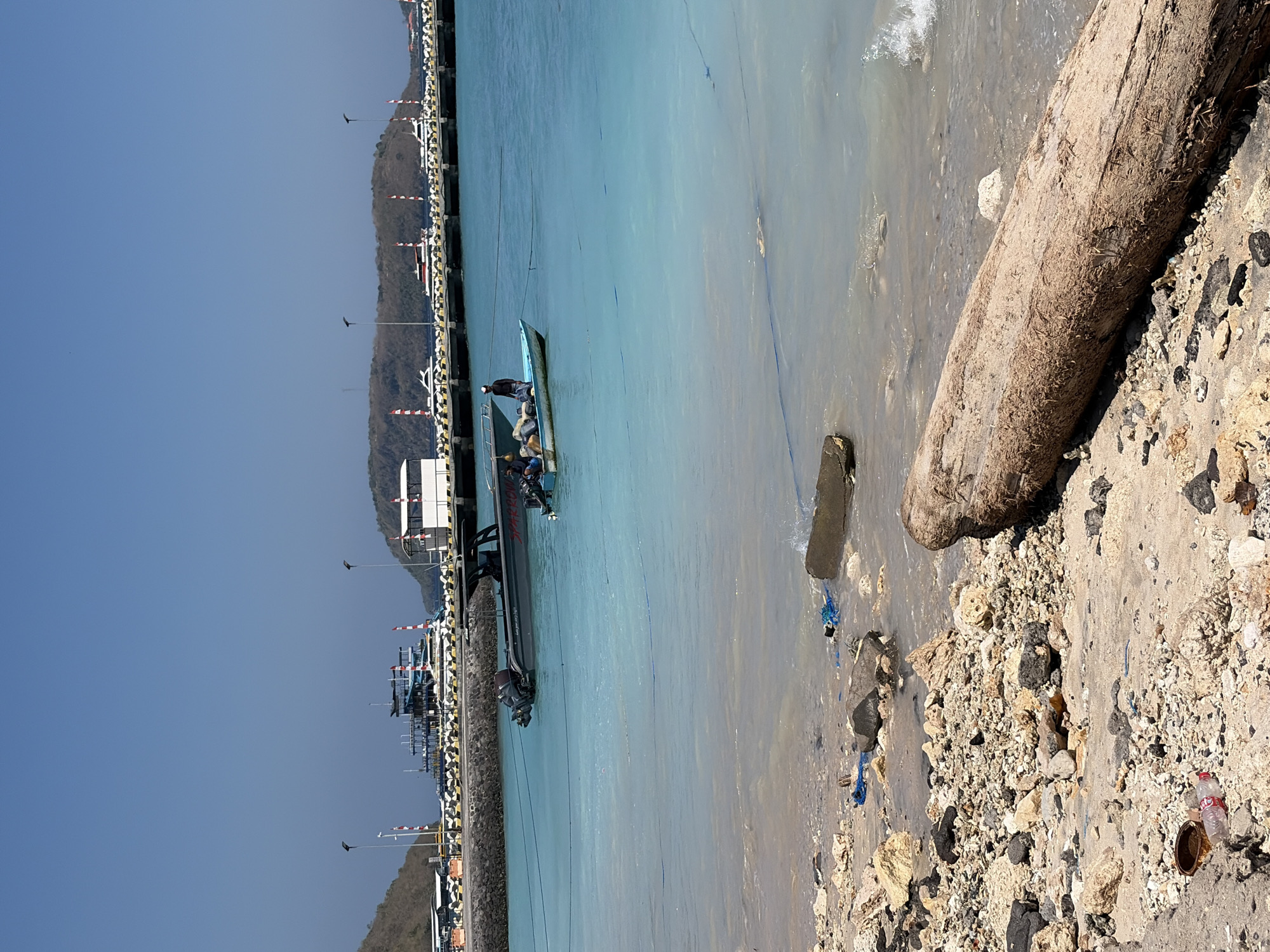 A scene of a coastline in Nusa Penida, Indonesia, depicts a rocky and sandy shore with scattered debris, including plastic bottles and rope. The turquoise water is shallow near the shore, revealing its sandy bottom. In the mid-ground, a dark-colored speedboat with two outboard motors is moored. Further out, a small, light-colored boat with at least two individuals aboard is also visible. In the background, a long pier stretches out, lined with several white speedboats and a larger multi-deck vessel. The sky is clear and blue, and a hilly, vegetated landscape forms the backdrop.