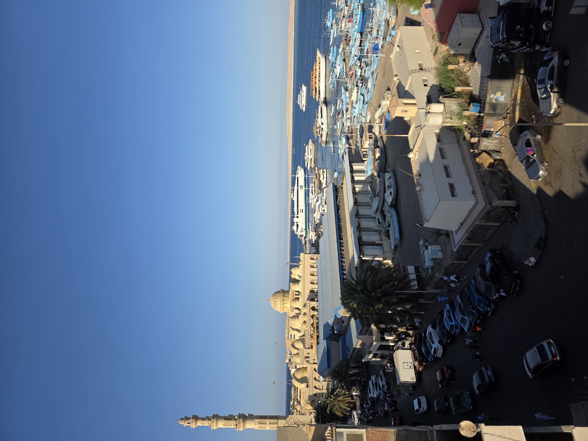 This is an aerial, eye-level, full shot of a port and mosque in Hurghada, Egypt, on a clear, sunny day. The scene is divided into a foreground of a street with parked cars and people, a middle ground of buildings and a mosque, and a background of a harbor filled with boats and the sea.
In the foreground, a street is lined with parked cars, including a black truck, several sedans, and some smaller vehicles. People are scattered along the street, some walking, others standing in groups. The street is paved and appears to be somewhat uneven. There are some buildings on the left and right sides of the street, one of which has a reddish-pink facade.
The middle ground is dominated by a large, light-colored mosque with multiple domes and a tall, slender minaret. The mosque is surrounded by other buildings, some of which appear to be commercial or administrative in nature, with flat roofs and windows. There are palm trees and other vegetation visible between the buildings. A large body of water, the sea, is visible beyond the buildings.
The background is a busy harbor filled with numerous boats of various sizes, from small fishing vessels to larger yachts and ferries. The water is a deep blue, and the horizon is marked by a strip of sandy land. The sky is a clear, bright blue. The overall impression is of a bustling port town with a significant religious landmark.