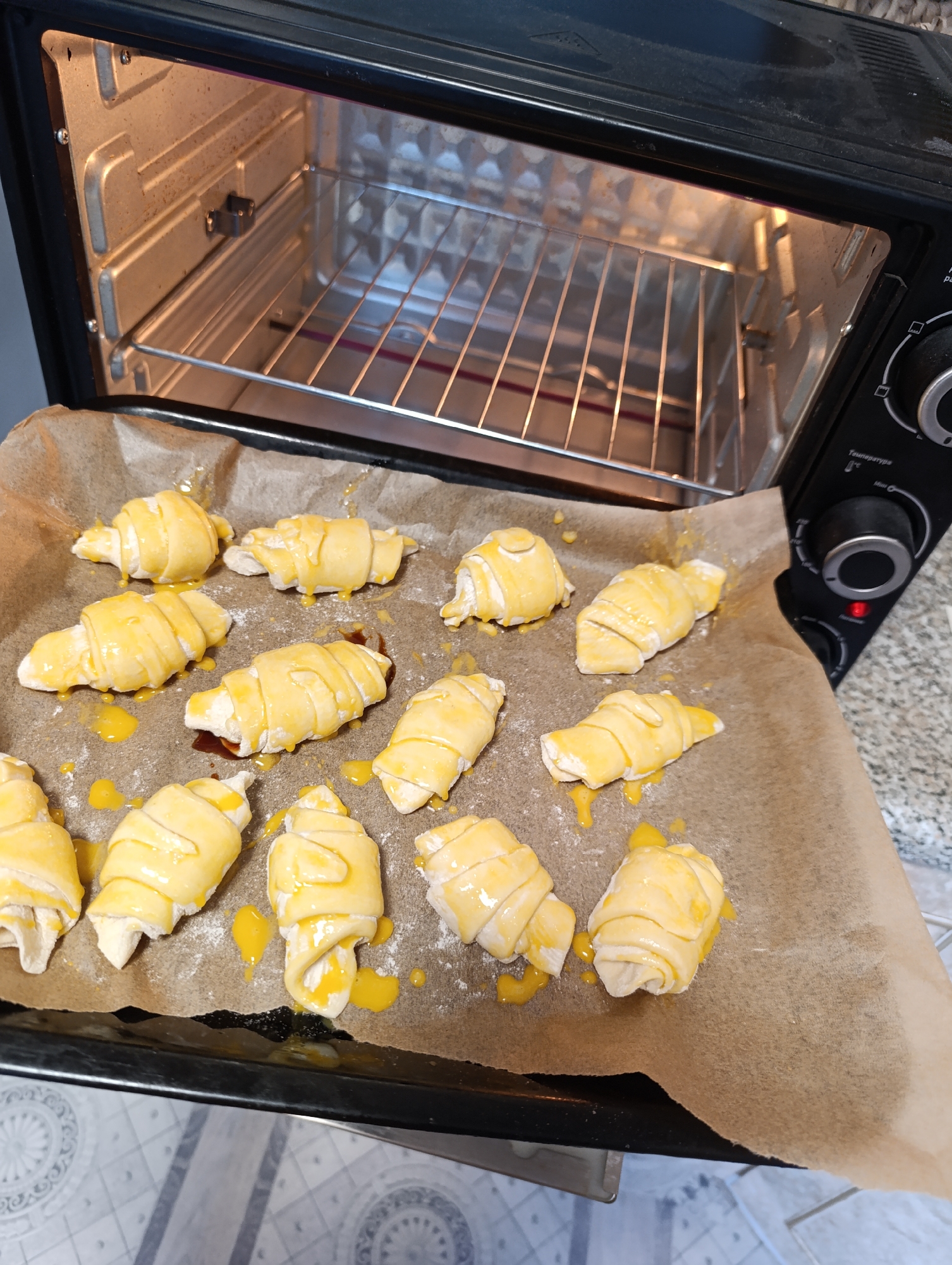An indoor, eye-level shot shows a black toaster oven with a baking tray of uncooked crescent rolls on parchment paper. The crescent rolls are coated in an egg wash and some are leaking filling. The baking tray is positioned in front of the open oven door, with the oven rack visible inside. The oven appears to be clean and functional, with a light on inside. The background consists of a kitchen counter and floor, with a blurred pattern. The scene is lit by overhead lighting, creating a warm and inviting atmosphere.