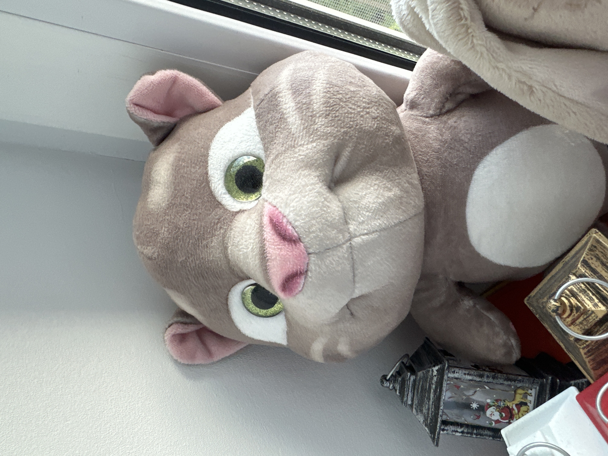A close-up, sideways shot shows a light brown plush cat toy with large, green eyes and pink ears. The toy is resting on a white surface, possibly a windowsill, next to a window with a slightly blurred view of greenery outside. The setting suggests a domestic environment. To the right of the cat toy are some decorative items, including a small, dark lantern with a Christmas scene visible inside, featuring Santa Claus and a reindeer, and a metallic gold lantern with a ring handle. A white and red object, possibly another lantern, is also partially visible. The lighting is bright, indicating daytime. There is no discernible text in the image. The plush toy's expression is neutral, and the overall scene is still, with no activity taking place. The location context provided is Zelenodol'sk, Russia.