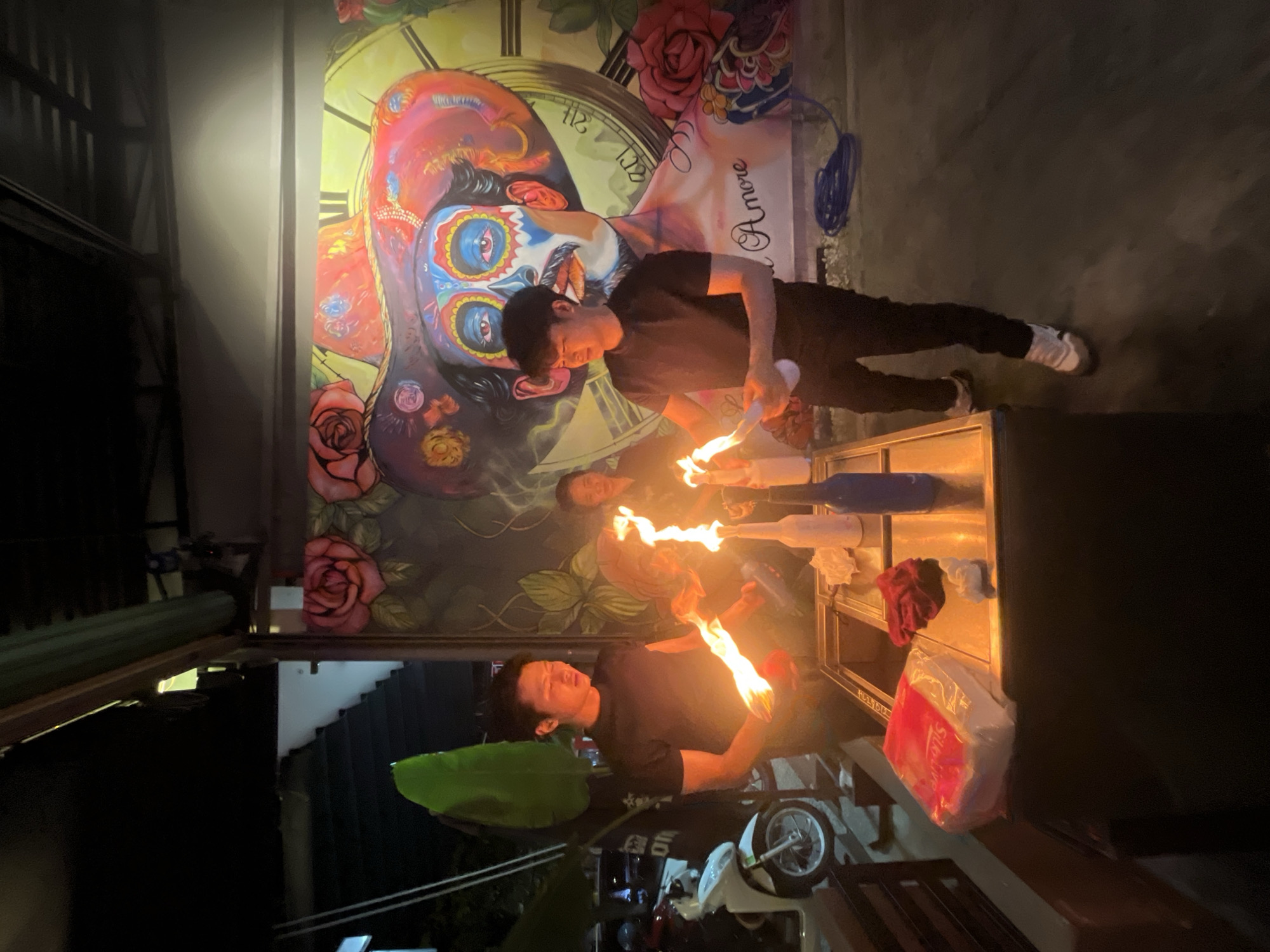 The image depicts an outdoor scene at night, likely in Đà Nẵng, Vietnam, where three individuals are engaged in a fire-juggling performance. The background features a large, vibrant mural of a person adorned with colorful face paint, reminiscent of Dia de los Muertos imagery, with roses and a clock face integrated into the design.
Two young men in black t-shirts and dark pants are the primary focus. The man on the left is holding a flaming object in his left hand, with his right hand raised. The man on the right is holding a flaming object in his left hand and appears to be in the process of juggling or demonstrating. Both are illuminated by the flames they are handling. A third person, a woman, stands behind them, partially obscured by the flames and shadows, observing the performance.
In the foreground, a table or counter holds various items, including a package of tissues, a red cloth, and two bottles, one white and one blue. The ground surface is dark and appears to be paved. A scooter is visible in the background to the left of the frame. The overall atmosphere suggests a casual, possibly entertainment-focused venue.