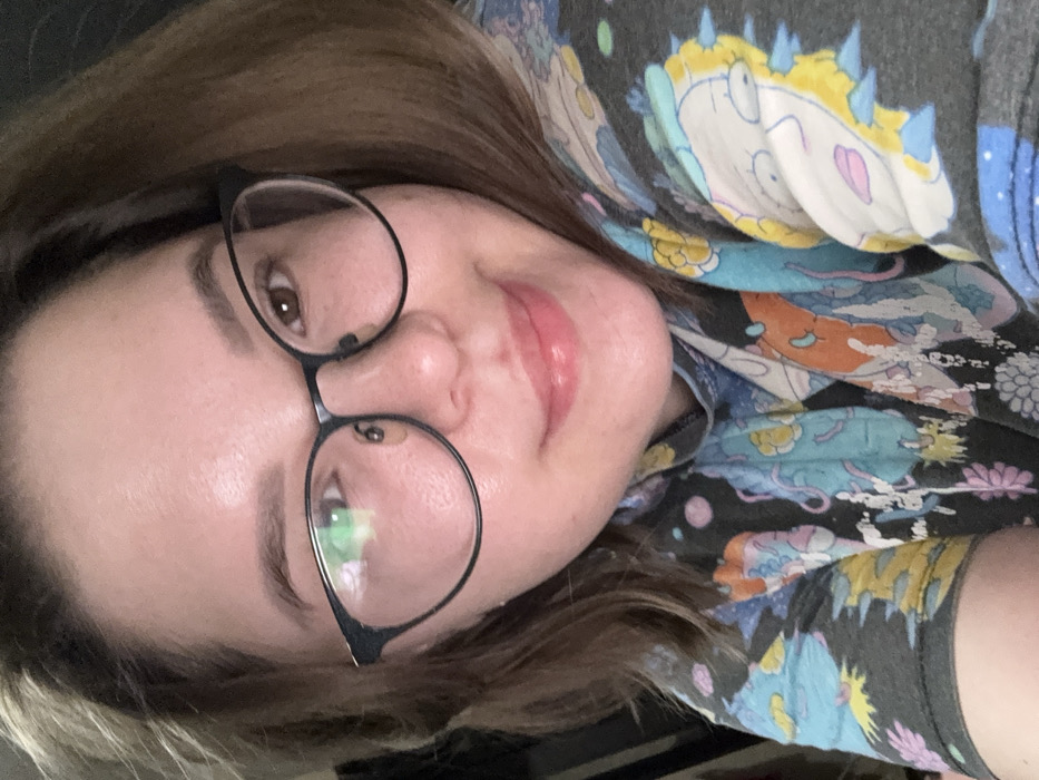 The scene depicts a close-up, chest-up portrait of an adult female located indoors in Kazan, Russia. The individual has light skin, brown shoulder-length hair, and brown eyes. She wears black-framed eyeglasses, with a visible reflection of a light source on the left lens. Her lips are slightly parted in a closed-mouth smile, exhibiting a glossy appearance. She is dressed in a dark gray short-sleeved top, likely a t-shirt, featuring a colorful, abstract pattern composed of light blue, yellow, orange, pink, and white stylized elements, including what appears to be a character's head or face on the right shoulder area. The background is dark and uniformly out of focus, providing no specific environmental details beyond an implied indoor setting. The subject is looking directly towards the camera.