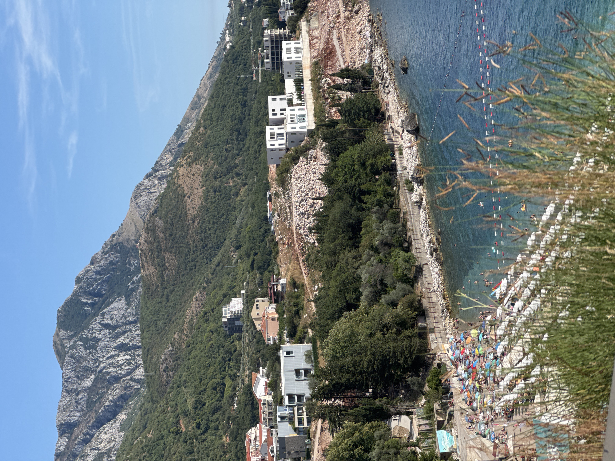 This image captures a sun-drenched scene in Kamenovo, Montenegro, featuring a bustling beach and modern architecture set against a backdrop of rugged mountains. The foreground is dominated by a stretch of beach covered with numerous colorful umbrellas and beach chairs, indicating a significant number of people enjoying the sunny weather. Many individuals are visible, some lounging on chairs, others walking or standing. The sea is a clear blue, with some people swimming and others relaxing on inflatable floats. Protective buoys mark swimming areas in the water.
Behind the beach, a paved walkway runs along the coastline, bordered by a stone wall. Adjacent to the walkway, a scattering of rocks and vegetation line the water's edge. Further inland, a mix of contemporary buildings and older structures are situated on a hillside. Prominent among these are stark white, modern apartment complexes with geometric designs, some appearing to be under construction with visible scaffolding and heavy machinery like cranes in the distance. Lower down the slope, residential buildings with red and orange tile roofs are nestled amongst greenery. The upper slopes of the mountains are covered in dense green foliage, transitioning to rocky, barren peaks at the highest elevations, all under a clear blue sky. The overall scene depicts a vibrant coastal resort area combining leisure activities with ongoing development.