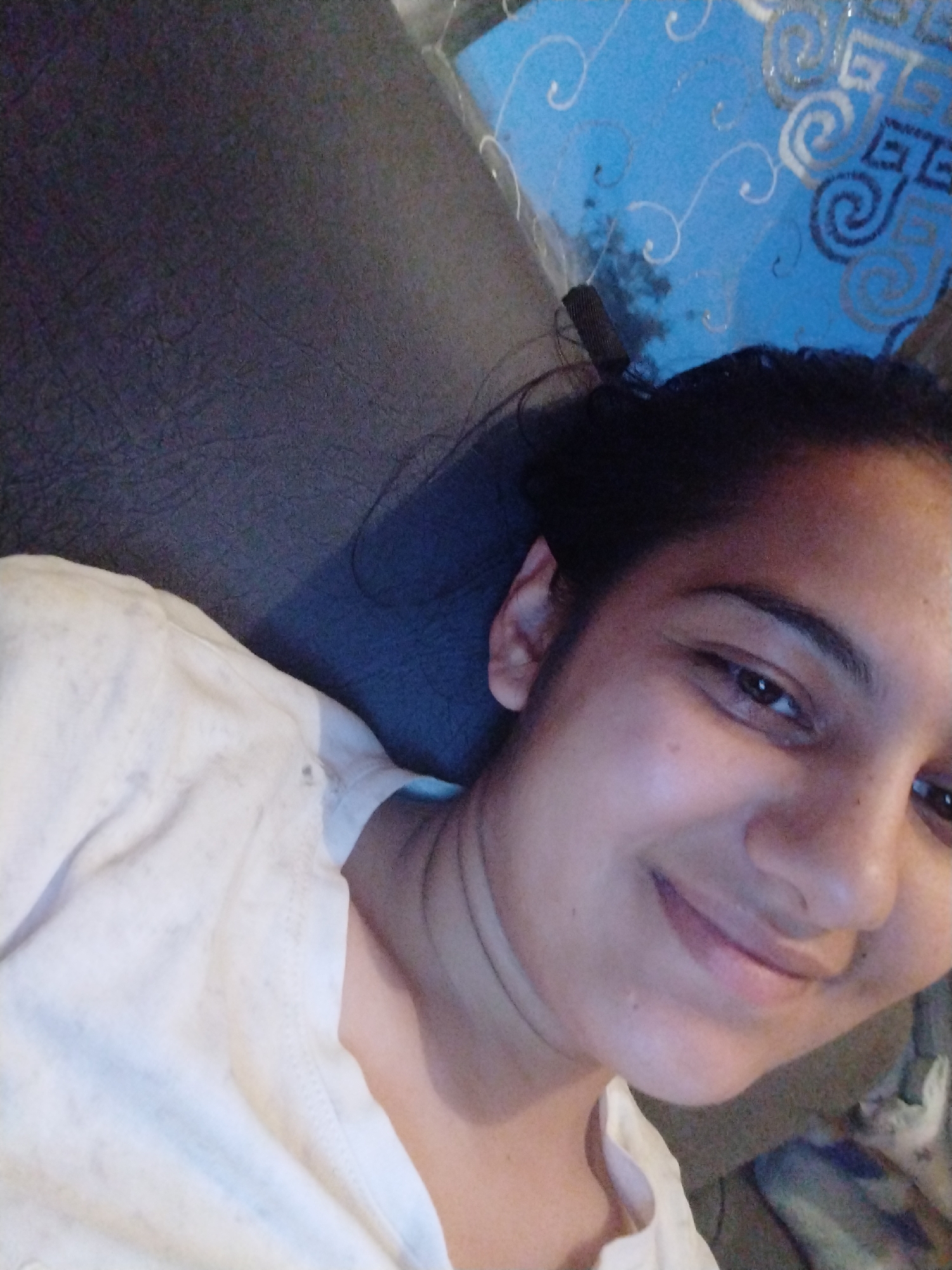 A person with dark hair, pulled back, and wearing a white shirt is shown in a close-up, selfie-style image. The person is lying on what appears to be a grey couch or cushioned surface, with a blue patterned object visible in the background. The location is Unknown, Unknown.