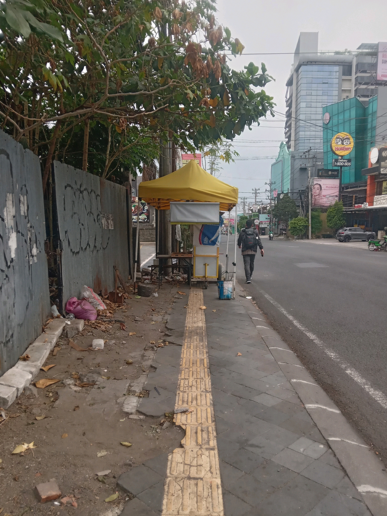This image depicts a street scene in Depok, Indonesia, focusing on a sidewalk adjacent to a busy road. A person with a backpack is walking away from the camera on the sidewalk. In the foreground, the sidewalk is partially damaged, with exposed soil and scattered debris including leaves and plastic bags. A yellow tented cart, possibly a food stall, is set up on the sidewalk next to the road, with a small table and a blue bucket. A metal fence with graffiti lines the left side of the sidewalk. Beyond the fence, trees with green and brown leaves are visible. Across the road, modern buildings with glass facades and billboards stand, some of them illuminated. Several vehicles, including cars and motorcycles, are present on the road, indicating ongoing traffic. The sky is overcast.