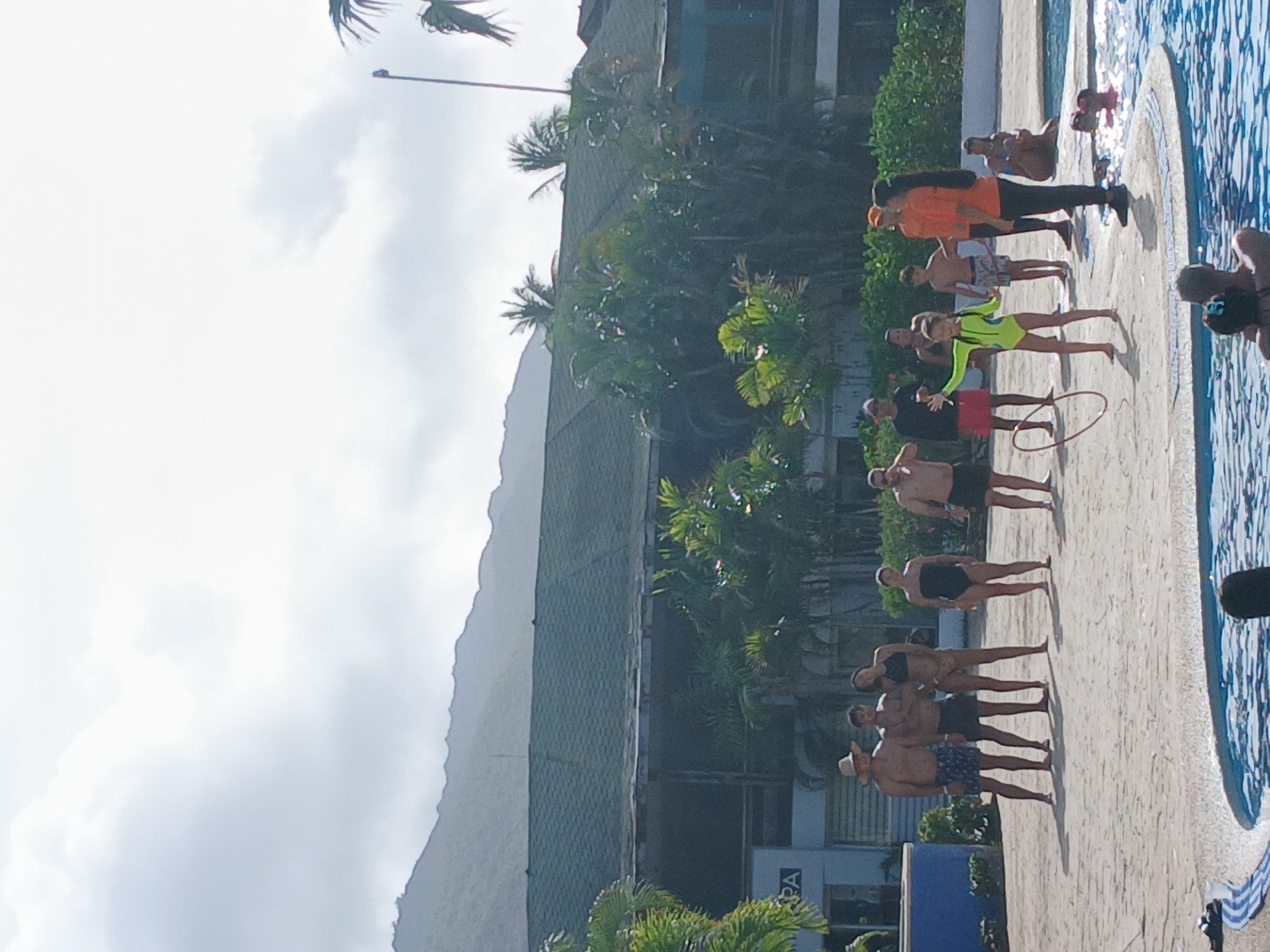 A group of people are standing near a pool in Playa El Agua, Venezuela. There are several adults in swimwear and casual clothes. Some are interacting with the water's edge, while others appear to be posing for a photo. One person is holding a hula hoop, and another is wearing an orange shirt and black pants. The environment includes tropical foliage, a large building with a dark roof, and a hazy mountain in the background. The sky is overcast.