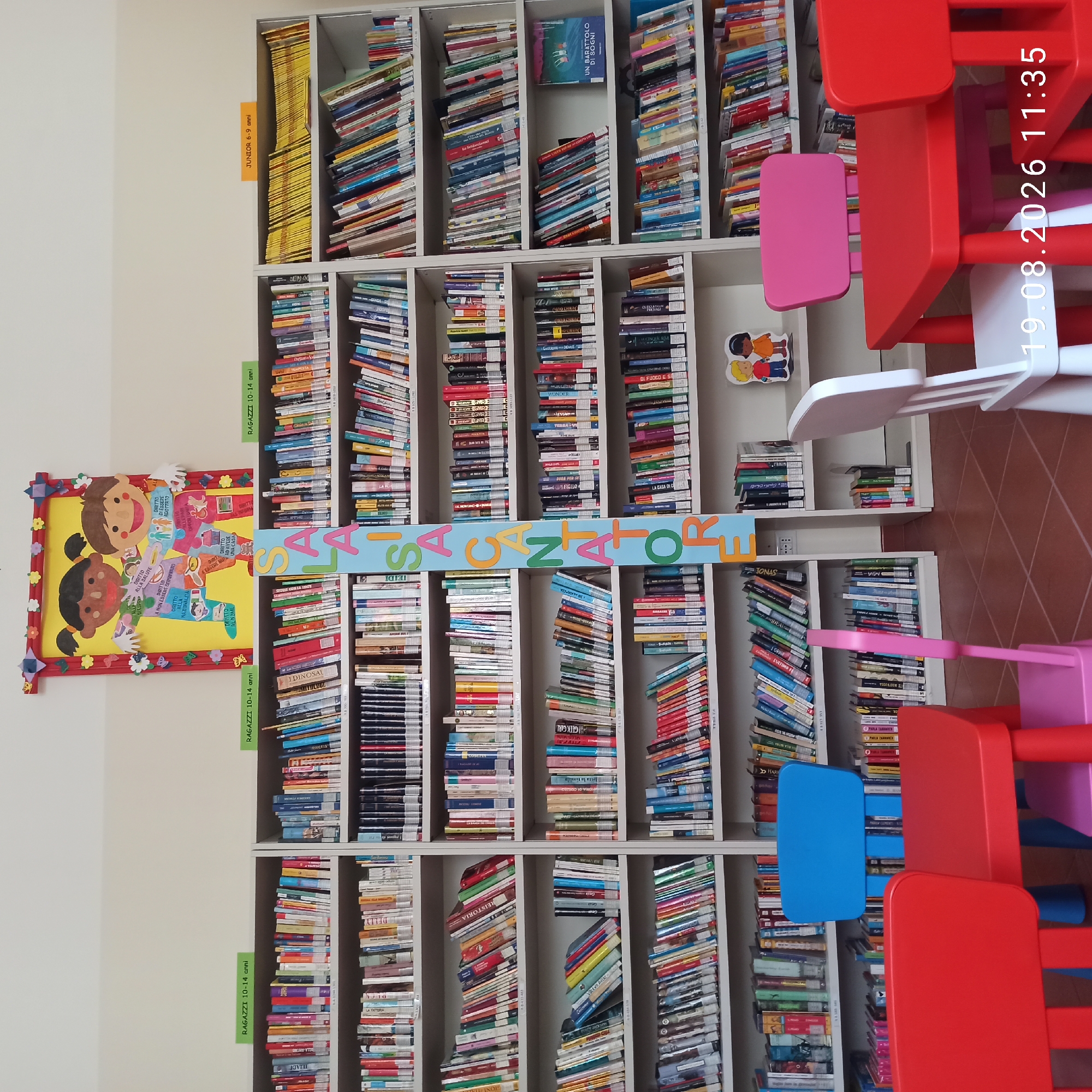 The image depicts the interior of a library, likely a children's section, located in Ruvo di Puglia, Italy. The scene is dominated by tall bookshelves filled with numerous books of varying colors and sizes. The shelves are organized by age groups, indicated by signs in Italian such as "RAGAZZI 10-14 anni" (Boys/Girls 10-14 years) and "JUNIOR 6-9 anni" (Junior 6-9 years). A large, colorful vertical sign with the letters "SALSI CANATORE" in bright, rounded characters is prominently displayed between two sections of bookshelves.
In the foreground, there are child-sized tables and chairs. A red table is positioned in the center, with a blue chair to its left and a pink chair to its right. A white chair is visible to the left of the red table. Another red chair is partially visible on the right. The floor is tiled with a pattern of terracotta-colored squares.
On the top shelf of one of the bookshelves, a colorful cutout featuring two children, one with dark skin and the other with light skin, holding hands, is mounted on the wall. This cutout also has text in Italian listing rights such as "DIRITTO ALLA SALUTE" (Right to Health) and "DIRITTO DI ESSERE ADOTTATO" (Right to be Adopted).
The overall environment suggests a well-equipped and inviting space for young readers. The lighting appears to be artificial, casting a neutral illumination on the scene. No people are actively engaged in any discernible actions in the image.