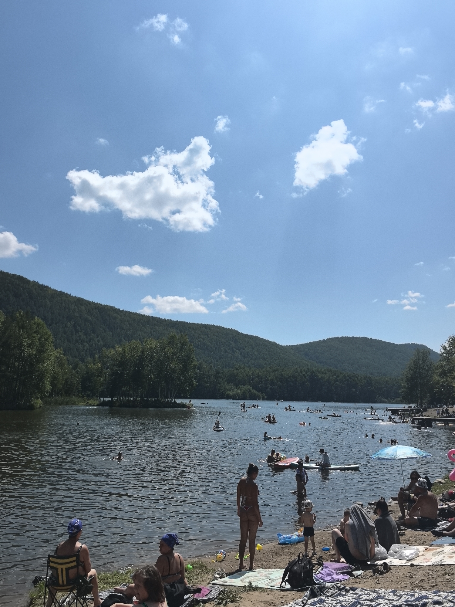 The image depicts a crowded summer day at a lake, likely the Solnechnyi reservoir in Solnechnyi, Russia, given the context. The sky is a clear blue with scattered white clouds. The foreground shows a sandy beach area where several people are relaxing. Some are seated on towels or beach chairs, while others are standing. There are various beach items present, including bags, towels, and what appear to be toys.
The lake itself is filled with people engaged in water activities. Many are on paddleboards, some are swimming, and a few are on inflatable rafts. The water is choppy with sunlight reflecting off its surface. In the background, a densely forested hillside rises from the edge of the lake, suggesting a natural, possibly park-like setting. The overall atmosphere is one of a popular recreational spot during warm weather.