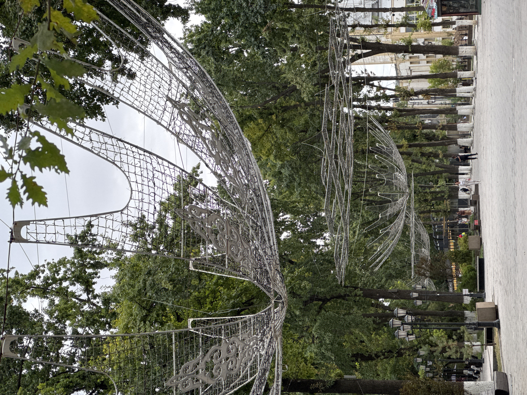 An outdoor shot captures a pedestrian walkway in Shymkent, Kazakhstan, with an overhead canopy of decorative metal structures adorned with lights and intricate patterns. These structures, resembling large, inverted flower petals or wings, are suspended between trees and supported by unseen frameworks. The walkway is paved with light-colored, rectangular stones. Along the sides of the path, numerous tall trees with dark bark, many with their lower trunks painted white, create a canopy of green foliage. Several lampposts with multiple ornate lights are positioned along the walkway. In the background, buildings with light-colored facades are visible, and a few people are seen walking or standing. The overall atmosphere is that of a public park or square, likely a popular urban space.