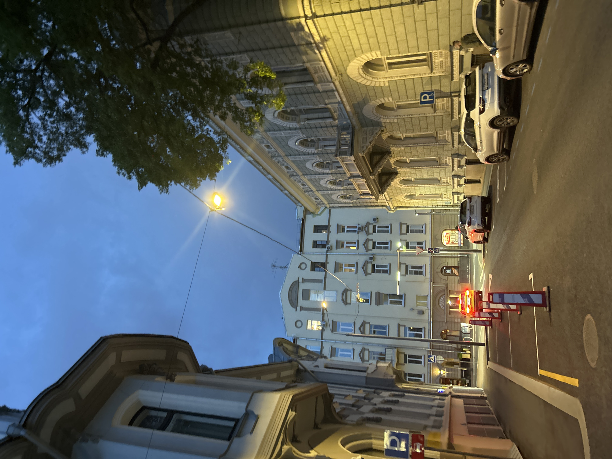 Captured at an oblique angle, the image presents a city street scene at dusk. The sky is transitioning from daylight to night, and illuminated streetlights cast a warm glow on the architecture and pavement. Buildings with varying styles line the street; some exhibit classic architectural details, while others are more modern apartment buildings. Cars are parked along the right side of the street. Roadwork is indicated by barricades with flashing red lights. No people are visible in the immediate foreground. A tree branch is visible in the upper left. The location is Unknown, Unknown.