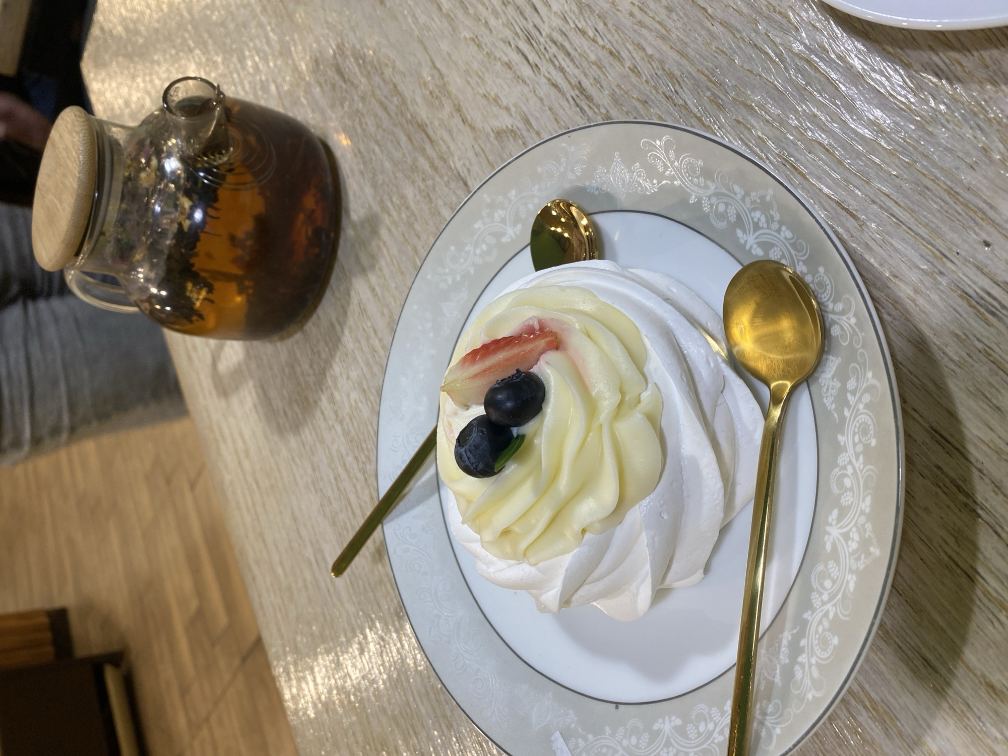 The scene captures an indoor tabletop arrangement, consistent with a cafe or restaurant environment, specified as being in Izhevsk, Russia, though no specific visual identifiers for the city are present within the image frame. The primary subject is a dessert, approximately 10-12 cm in diameter, centered on a white plate with a light brown rim featuring intricate white floral and scrollwork patterns. The dessert consists of a white meringue base topped with piped pale yellow cream or custard. Garnish includes two intact dark blueberries, one vertically sliced red strawberry, and a single small green leaf. Two small, reflective golden spoons are positioned; one rests on the plate to the lower right of the dessert, and a second is partially visible behind the dessert, extending from the upper left. To the upper right, a clear glass teapot with a light wooden lid contains a dark amber liquid with visible steeped tea leaves or herbs. The table surface is light-colored wood or laminate with a discernible grain pattern. In the blurred background, a person's left leg, clothed in grey fabric, is partially visible behind the teapot. Further back, a light-toned wooden floor with rectangular planks is visible.