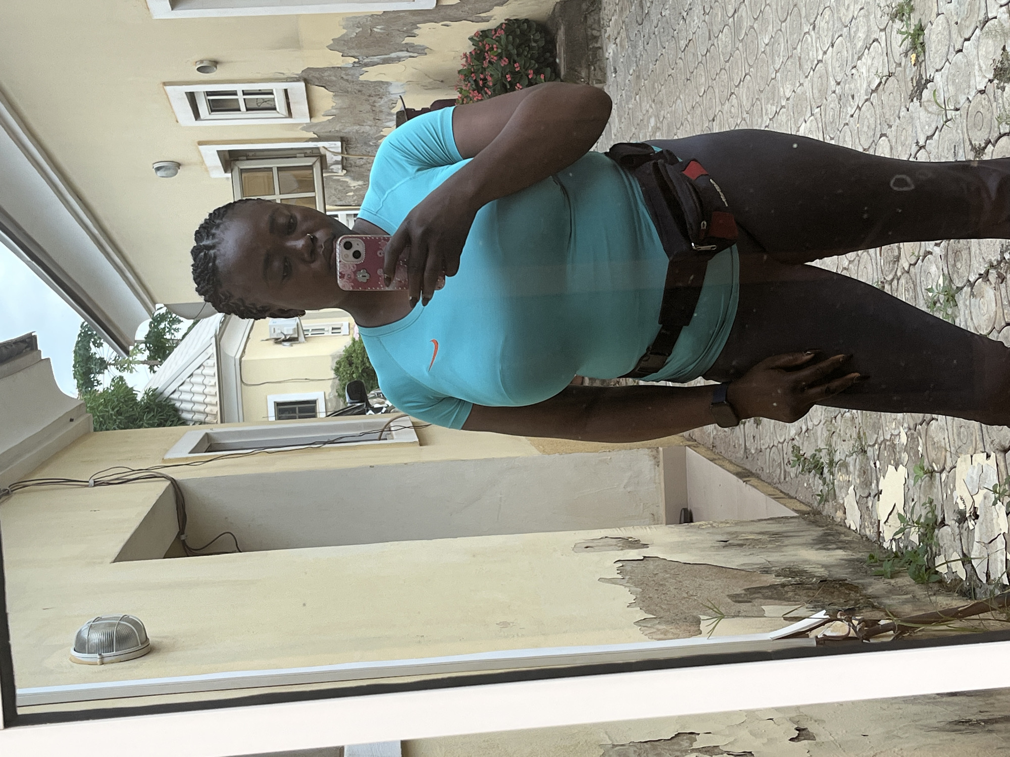 A full-body selfie taken in an outdoor courtyard in Abuja, Nigeria, features a woman of color wearing a turquoise athletic t-shirt and dark leggings. She has her hair braided and is holding a pink and white patterned smartphone to take the photo. A black fanny pack is cinched around her waist. The background consists of pale yellow buildings with weathered exteriors and visible cracks and peeling paint. Cobblestone paving covers the ground, with small green plants growing in patches. A wall with an outdoor light fixture is visible on the left. Another building with white-framed windows and a tiled roof is partially visible in the background. The lighting suggests an overcast day.