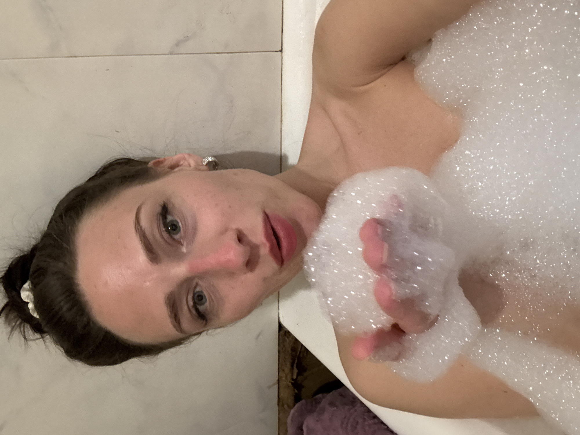 A woman is in a bathtub filled with bubbles. She is partially submerged, with her face and one arm visible. The background appears to be a tiled bathroom. There is no indication of the location being Saint Petersburg, Russia.