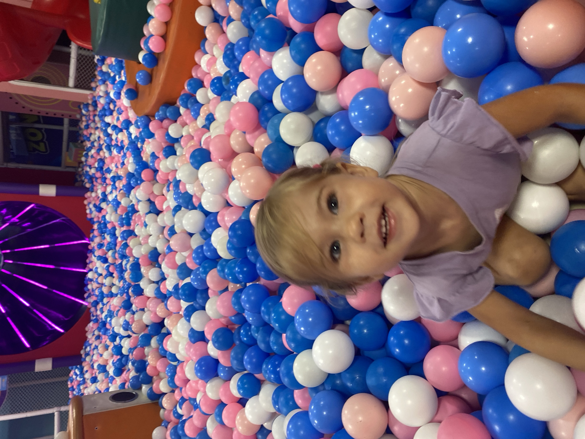 A young girl with blonde hair and blue eyes is pictured in a ball pit filled with pink, blue, and white balls. She is wearing a purple t-shirt with ruffled sleeves and is smiling at the camera. The ball pit appears to be part of a play area, with colorful structures and lights visible in the background. The overall atmosphere suggests a fun and playful environment. The location of Phu Thuy, Vietnam, is not discernable from the image itself.