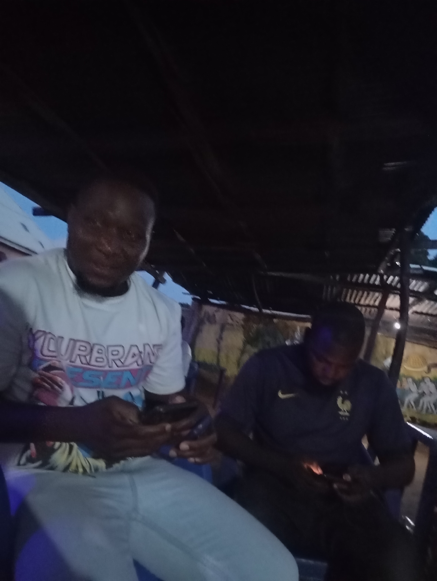 In Shendam, Nigeria, two men are seated under a thatched roof structure at dusk. The man on the left, in a white t-shirt with text reading "YURBRAN ESEN" and light blue jeans, is looking at his phone. The man on the right, wearing a dark blue shirt with a Nike logo and a crest, is also looking at his phone. The environment appears to be an outdoor seating area, possibly a bar or restaurant, with dimly lit surroundings and visible wooden beams overhead.