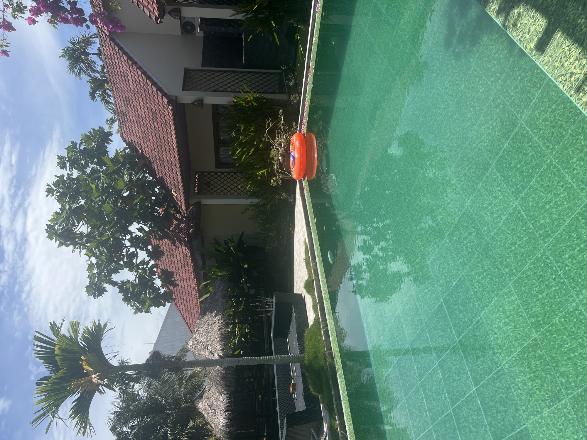 A swimming pool with turquoise water is in the foreground, tiled with small green squares. An orange inflatable ring floats on the surface of the water. In the middle ground, there is a black sofa set on a paved area, surrounded by grass and tropical plants. Behind the sofa, there are two thatched-roof huts. Further back, a building with a red tiled roof and cream-colored walls is visible, with palm trees and other greenery in the background. The sky is blue with scattered white clouds. The scene appears to be a resort or hotel in Hoi An, Vietnam.