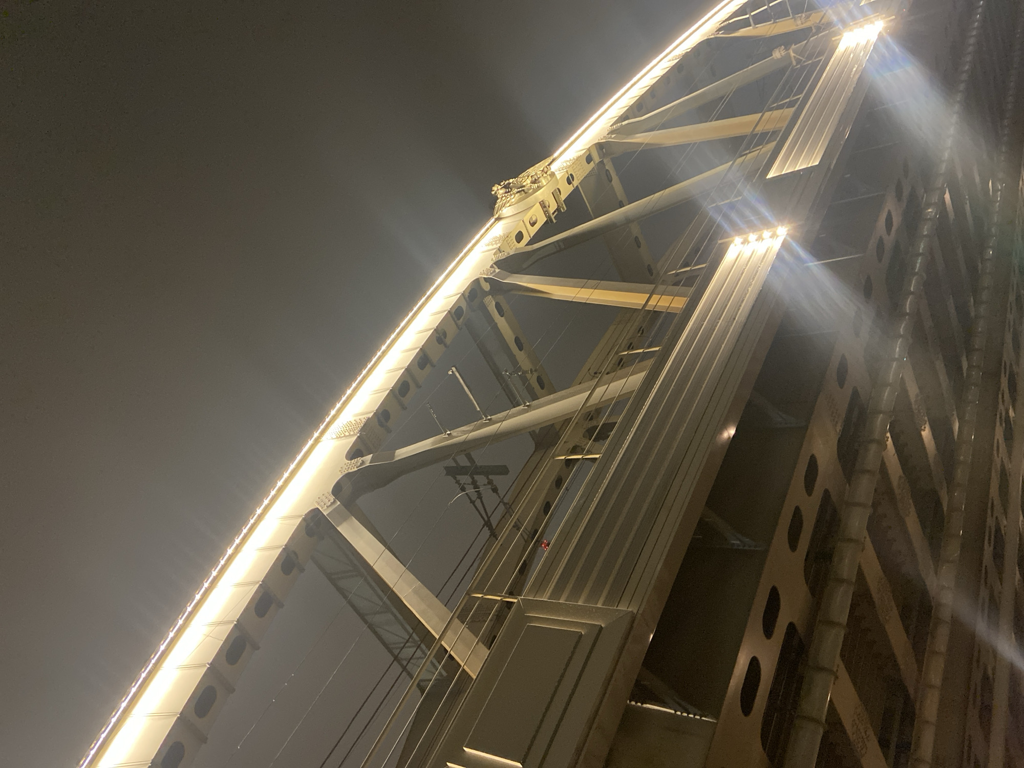 The image captures a close-up, low-angle view of a bridge structure at night, likely in Moscow, Russia, given the architectural style and lighting. The structure is composed of metal beams, cables, and support columns, illuminated by a string of lights tracing its arched form. The ambient light is diffused, creating a hazy or foggy atmosphere. The perspective emphasizes the height and complexity of the bridge's construction.