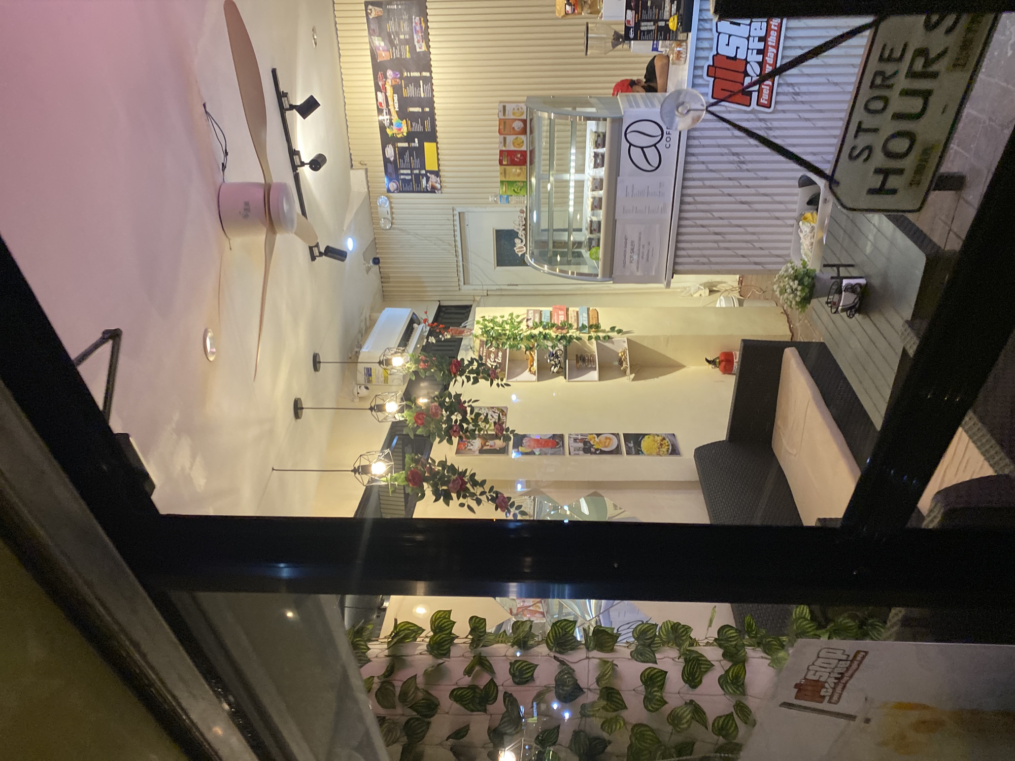 This image captures the interior of a Pitstop Coffee shop, specifically located in Matina, Davao City, Philippines, viewed through a glass storefront at night. The brightly lit cafe interior contrasts with the dark exterior, with reflections partially visible on the glass.
The environment features light-colored, vertically slatted walls. Artificial green leafy vines are draped vertically along a wall on the left, and clusters of artificial red roses are suspended from the ceiling, intertwined with modern cage-style pendant lights. A large, three-bladed ceiling fan is centrally mounted, with additional black track lighting fixtures directed towards the seating area.
Seating comprises dark-colored, possibly wicker or rattan, sofa benches equipped with light-colored cushions. A dark, rectangular table in the foreground holds a small white potted plant and a metal tray containing miscellaneous items. A "STORE HOURS" sign hangs vertically in the lower right foreground.
In the midground, a glass display case filled with various food items (likely pastries or desserts) rests on a counter. The display case features a "COFFEE" logo with a coffee bean graphic, and a sign within it advertises "CHICKEN BAGUET FOR SALE!!!". Behind this, a menu board displaying various drinks and food items is affixed to the slatted wall. A partial "Pitstop Coffee" logo with the slogan "Fuel your day the right..." is visible on the right side of the counter. A red fire extinguisher is situated on the floor to the left.
One individual, wearing a red cap, is partially visible behind the counter on the right, appearing to be attending to the coffee preparation area. The scene depicts a quiet, inviting atmosphere within the coffee shop.