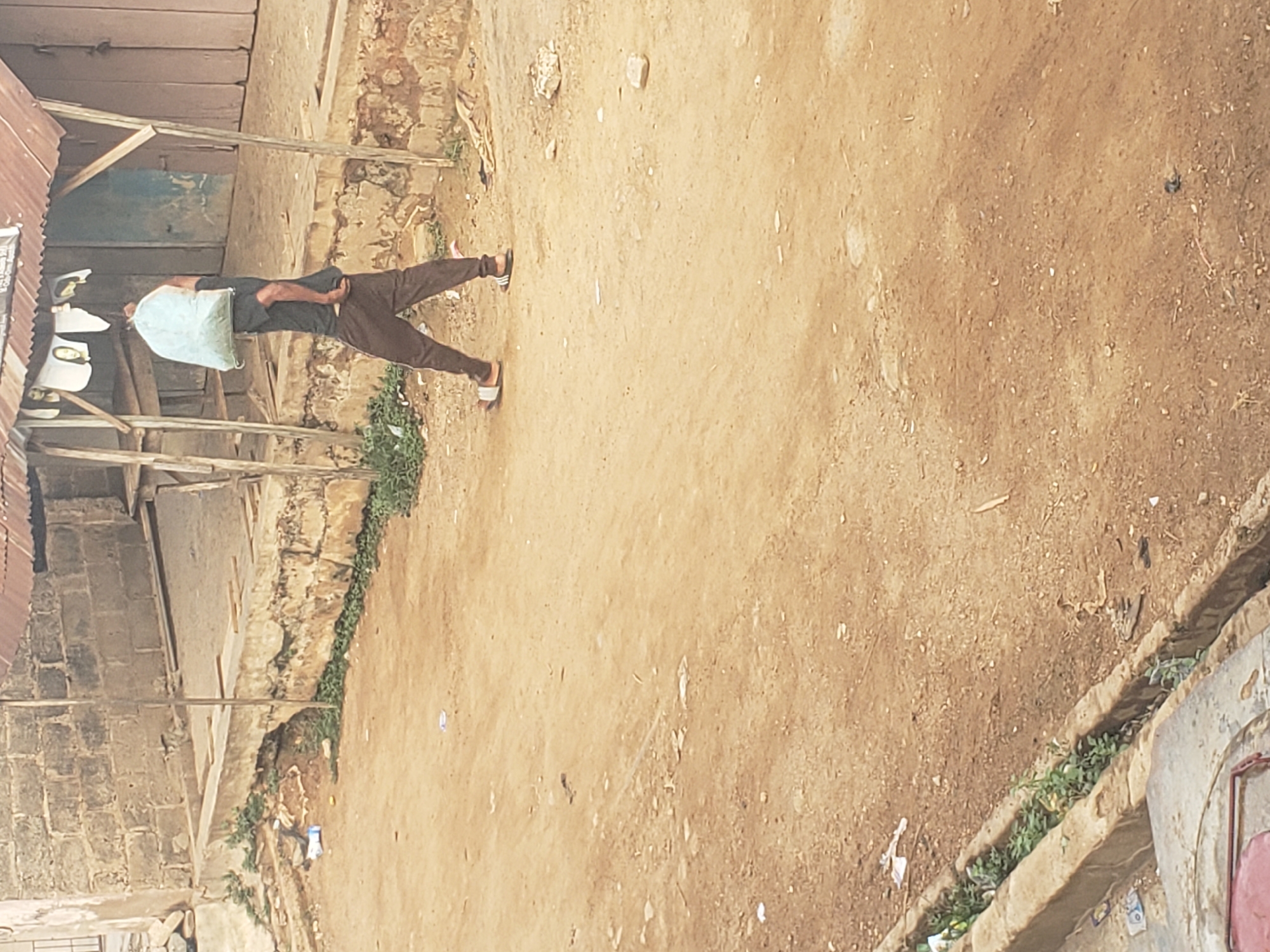 The image shows a man carrying a large light-colored bag over his shoulder, walking on a dirt ground in Alalubosa, Nigeria. He is wearing a dark shirt, brown pants, and sandals. The environment includes a building with a corrugated metal roof and brick walls on one side of the path, and a drainage ditch with vegetation on the other side. The ground is dry and dusty, with scattered debris.