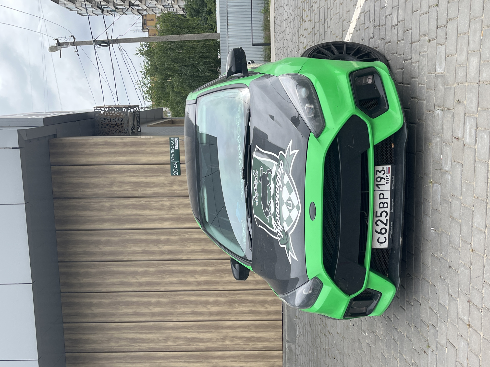 A bright green Ford Focus RS is parked on a cobblestone surface in front of a building with wooden siding. The car has a black hood with a large sticker featuring a green and white crest and checkered pattern. The license plate is visible and reads "C625BP 193 RUS". A street sign in the background indicates "URALSKAYA ULITSA 2046", which translates to "Uralskaya Street 2046". This location is in Krasnodar, Russia. The environment appears to be outdoors, with power lines and other buildings visible in the background. The sky is overcast.