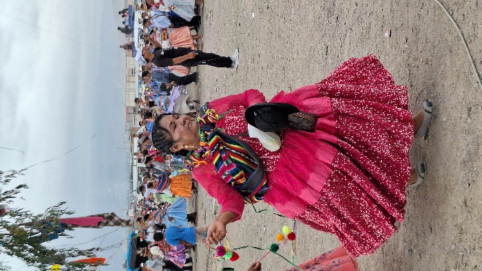 A woman in a vibrant pink dress, adorned with colorful pompoms, dances in a sandy outdoor setting. A striped shawl is draped over her shoulder, and a black hat rests on her back. In the background, a crowd gathers along a barrier, with a cloudy sky overhead. The composition captures a lively cultural celebration.