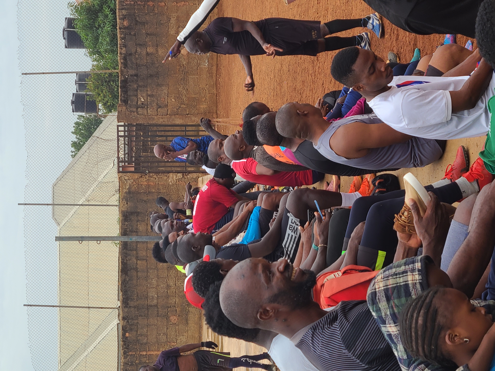 A group of approximately 30 people are sitting outdoors in an unidentified location within the city of Unknown, Unknown. They are seated on the ground, closely packed together. An individual dressed in black sportswear stands above them, appearing to instruct or address the group. The background consists of a brick wall, a fence, and greenery. In the upper-left corner, there are black water tanks above the fence. Some of the seated individuals are looking at their phones, while others are looking forward. The clothing worn varies, with some in sportswear and others in casual attire.