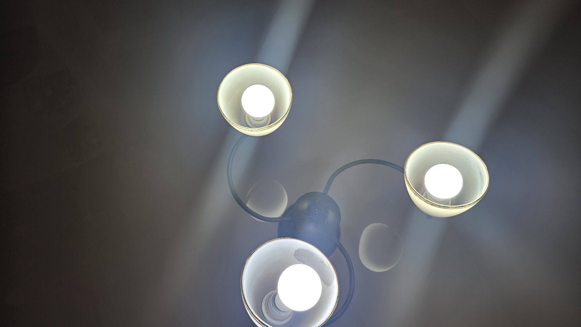 This is a close-up, top-down shot of a modern, three-bulb chandelier against a dark ceiling. The chandelier has a central black fixture with three curved arms, each holding a conical shade. The shades are cream-colored with a subtle gold trim. Inside each shade, a bright, white LED bulb illuminates. Light beams are visible, projecting outwards from the bulbs and casting shadows on the ceiling. The overall scene is clean and contemporary, with no people or other objects visible. The context of Chuy, Kyrgyzstan is not visually supported by the image content.
