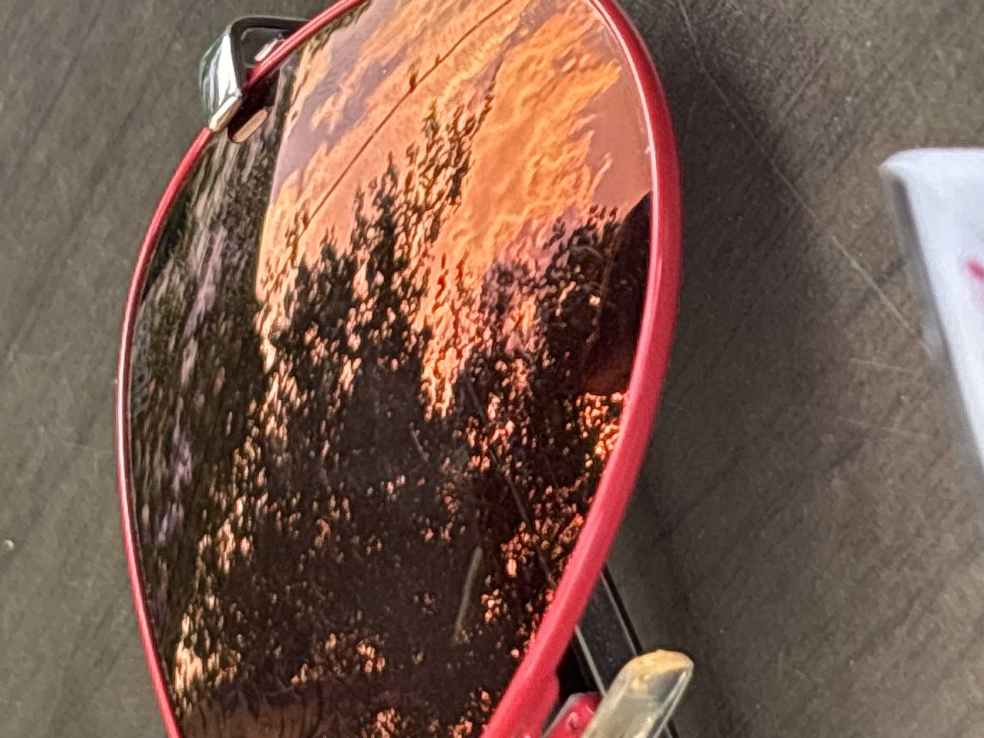 This is a close-up, slightly angled, full shot of red-rimmed sunglasses resting on a dark, textured surface, possibly a table. The lenses reflect a scene of trees silhouetted against a vibrant orange and yellow sunset sky. The reflection is detailed, showing individual leaves and branches, and the warm hues of the sky are prominent. The sunglasses appear to be outdoors, and the lighting suggests it is late afternoon or early evening. There are no people or active interactions visible in the image, and the location in Svoge, Bulgaria is not discernible from the visual information provided.