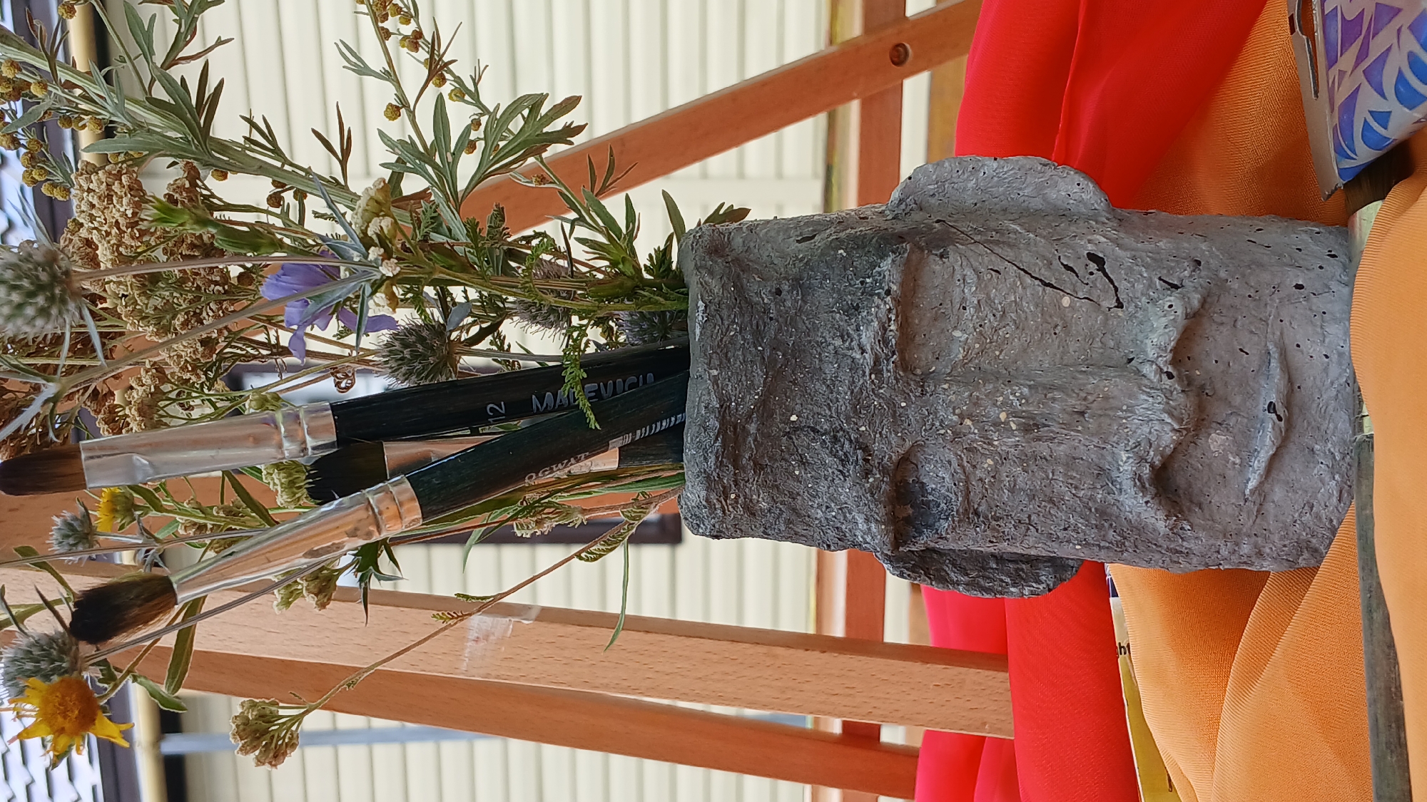 In the city of Derköl, Kazakhstan, a still life is arranged. A grey, concrete vase shaped like a stylized human head sits on a surface covered by orange and red fabric. The vase contains a bouquet of dried wildflowers and greenery, interspersed with three paintbrushes. Behind the vase, a wooden easel is visible. The background consists of a light-colored building with horizontal siding.