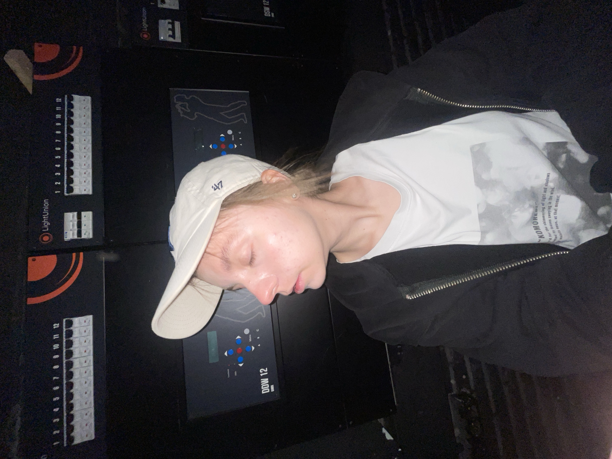 A fair-skinned individual wearing a white baseball cap, a white t-shirt, and a black zip-up jacket is lying down, with their face resting on a black, industrial control panel. The control panel features rows of switches, indicators, and a graphic of a human silhouette. The brand "LightUnion" is visible on the panel, along with labels for control functions like "DDW 12". The t-shirt has the word "KOMORE" printed on it, along with additional text about light and shadows. The background is dark and suggests an indoor setting. There is no evidence to suggest this image was taken in Moscow, Russia.