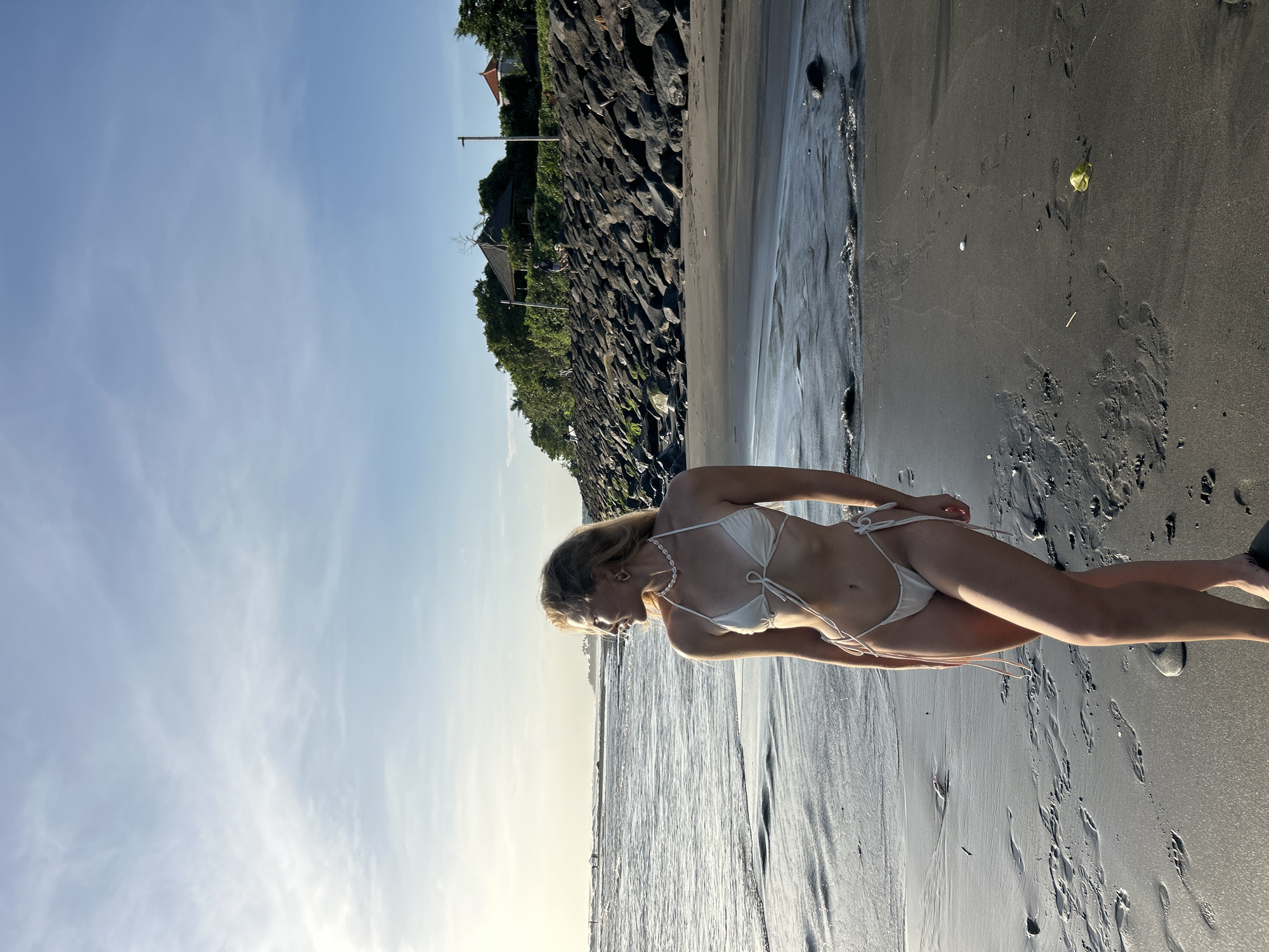 A young woman stands on a dark sand beach. She is wearing a white bikini and a pearl necklace. Behind her is a stone embankment with vegetation and a building at the top. The sky is blue. The photograph appears to be taken at the beach in Mengwi, Indonesia.