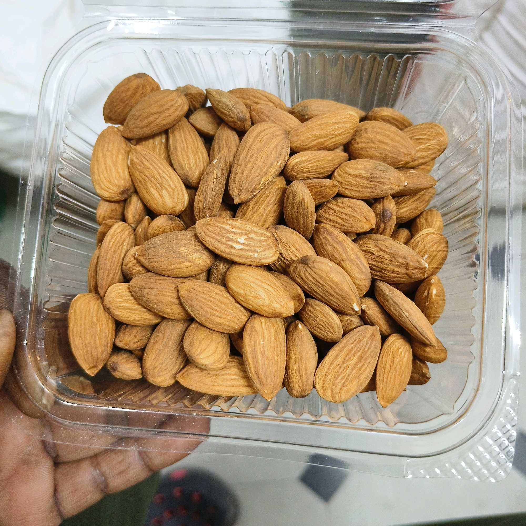 A close-up, eye-level, medium shot captures a clear plastic container filled with almonds. The container is situated on a white surface, possibly a countertop, with a person's hand holding the edge of the container on the left side of the frame. The almonds are light brown and appear to be whole and unpeeled. The plastic container has a ribbed texture along its lower edges and a smooth, curved upper rim. The background is blurred, but hints of a white surface and darker areas suggest an indoor environment. There are no discernible landmarks or specific details to indicate the location within Karimnagar, India. The scene is static, focusing solely on the presentation of the almonds in their packaging.