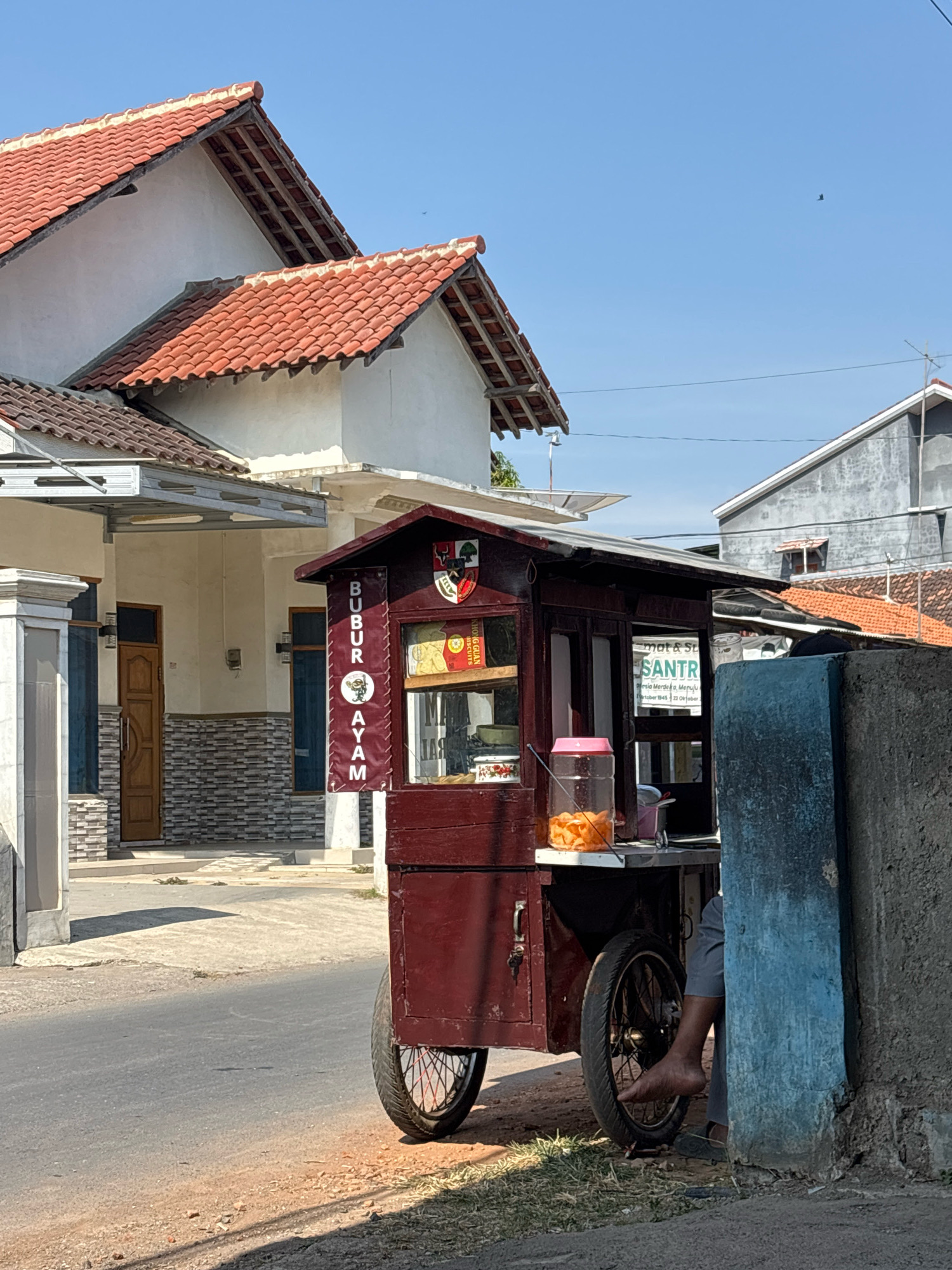The image shows a street scene in Tegal, Indonesia, dominated by a small, dark red mobile food cart. The cart is wheeled and appears to be stationary, positioned on the side of a paved road. It features a display window showcasing food items, with a banner above that reads "BUBUR AYAM" which translates to "Chicken Porridge." A small pink container filled with orange snacks is visible on the counter of the cart. A person, partially visible, is seated behind the cart, with their legs and bare foot extending out.
In the background, there are buildings with red tiled roofs and white walls, typical of Indonesian architecture. A blue wall is present on the right side of the image, bordering the food cart. The sky is clear blue, suggesting a sunny day. The overall impression is a slice of everyday life in a residential or commercial area of Tegal.