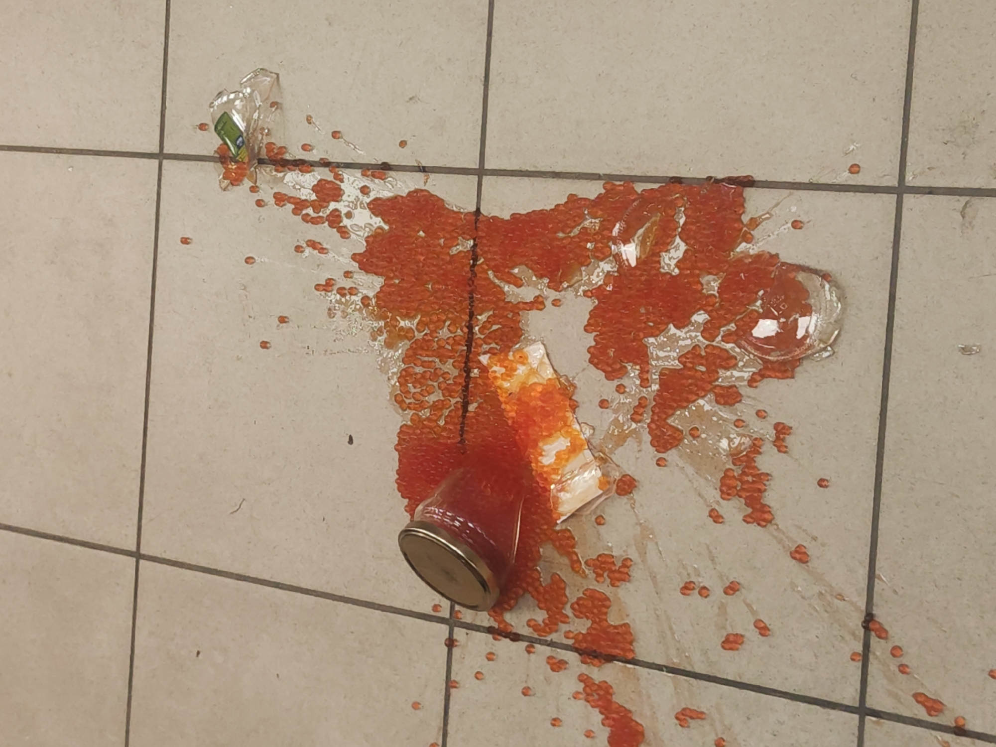 The image depicts a broken glass jar of orange fish roe (likely salmon roe) spilled on a light-colored, tiled floor. Fragments of glass from the broken jar are scattered around the spill. The jar's lid is visible near the base of the spill, and a torn piece of paper is partially submerged in the roe. The scene is indoors, with the tiled floor suggesting a commercial or institutional setting. There are no people visible in the image. The city of Pushkin, Russia, cannot be inferred from the visible elements.