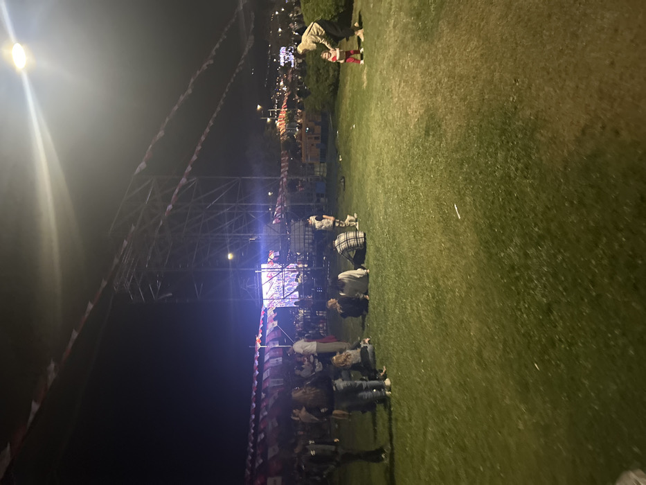 The image shows a group of people gathered in a grassy area in front of a stage, located at coordinates 38.433, 27.137, in İzmir, Türkiye. The stage is brightly lit, and there are several strings of red and white flags hanging above it. The people are standing and sitting on the grass, some are interacting, and one woman is walking with a young child. The scene is illuminated by streetlights and spotlights. There are trees and bushes surrounding the grassy area, and the background is a blurred image of buildings and trees. The image was taken at night.