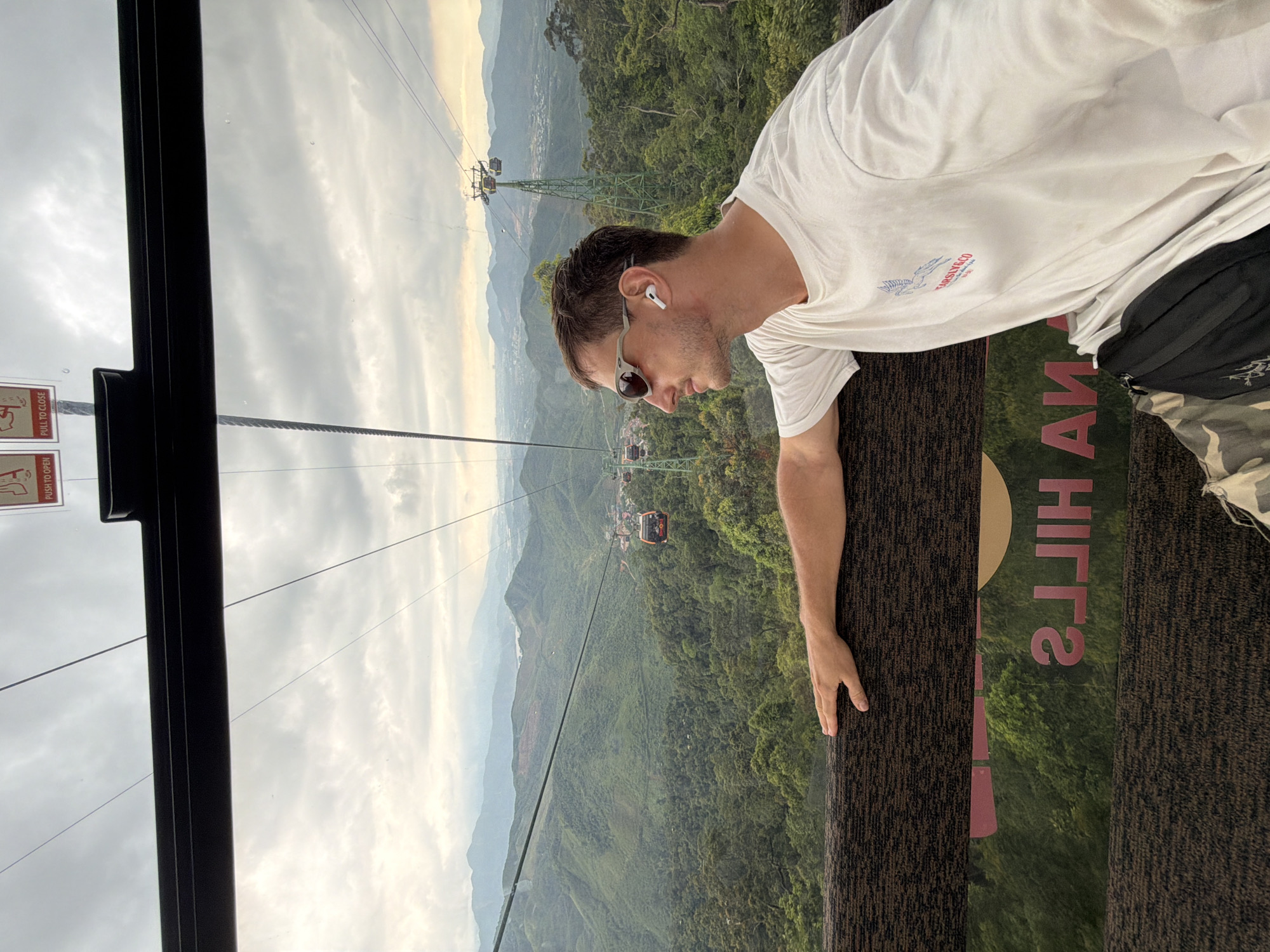 A medium, eye-level shot shows a man in a cable car ascending Bà Nà Hills in Đà Nẵng, Vietnam. The man is leaning against the side of the car, looking out. He is wearing sunglasses, an earbud, and a t-shirt. The cable car is suspended above a lush, green landscape. Other cable cars are visible in the distance. The sky is overcast. Part of the wall of the cable car is visible, including text reading "Hills."