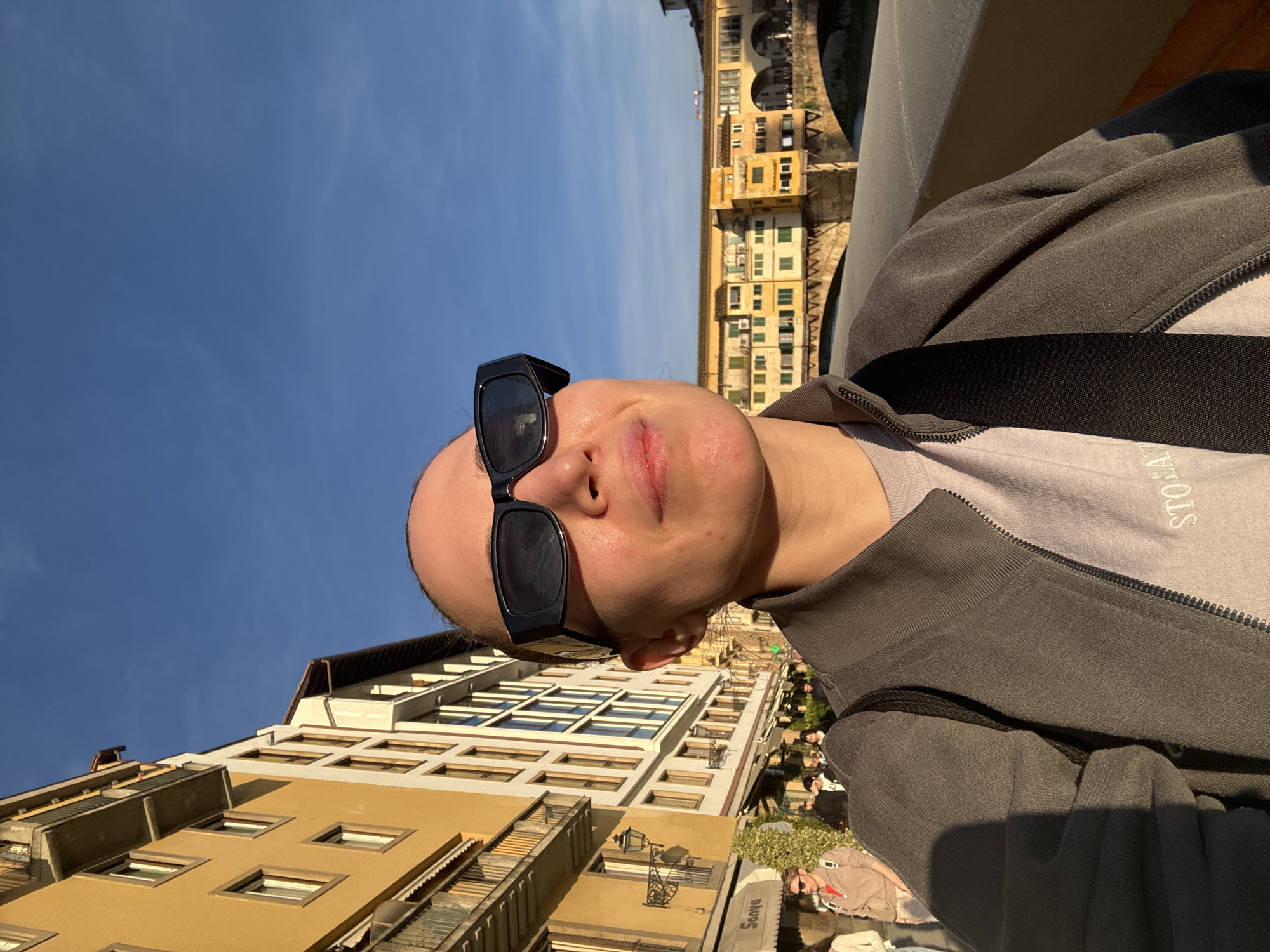 A medium-shot photograph shows a person wearing black sunglasses and a gray jacket against a backdrop of architecture and blue sky. The buildings appear to be old with varied colors from beige to brown, featuring windows and balconies. Some buildings appear to be adjacent to a river or other waterway. People can be seen near the lower right-hand side of the photo. The angle suggests the photo was taken looking upwards towards the sky and nearby buildings. Based on the architecture and setting, the location appears to be Florence, Italy, specifically near the Ponte Vecchio.