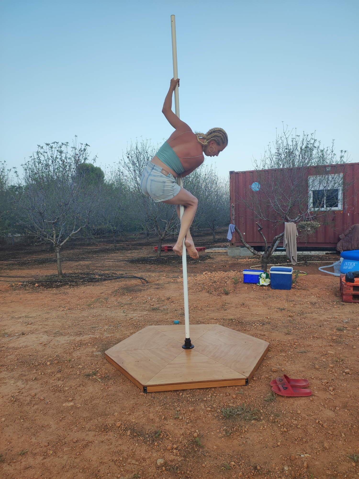 A woman with blonde braided hair is performing a pole dance on a portable stage outdoors. She is wearing a green halter top and denim shorts, with her legs wrapped around the pole. The setting appears to be a rural area with dry, reddish-brown earth, scattered rocks, and sparse vegetation. Several trees, some appearing to be fruit trees, are visible in the background, along with a red shipping container that has a window and a hanging towel. Near the stage, there are two blue coolers, a red pallet with a blue object on top, and a pair of red flip-flops. The sky is clear and light blue, suggesting it is either early morning or late evening. The image does not contain any specific visual cues that would indicate the location as Alcàsser, Spain.