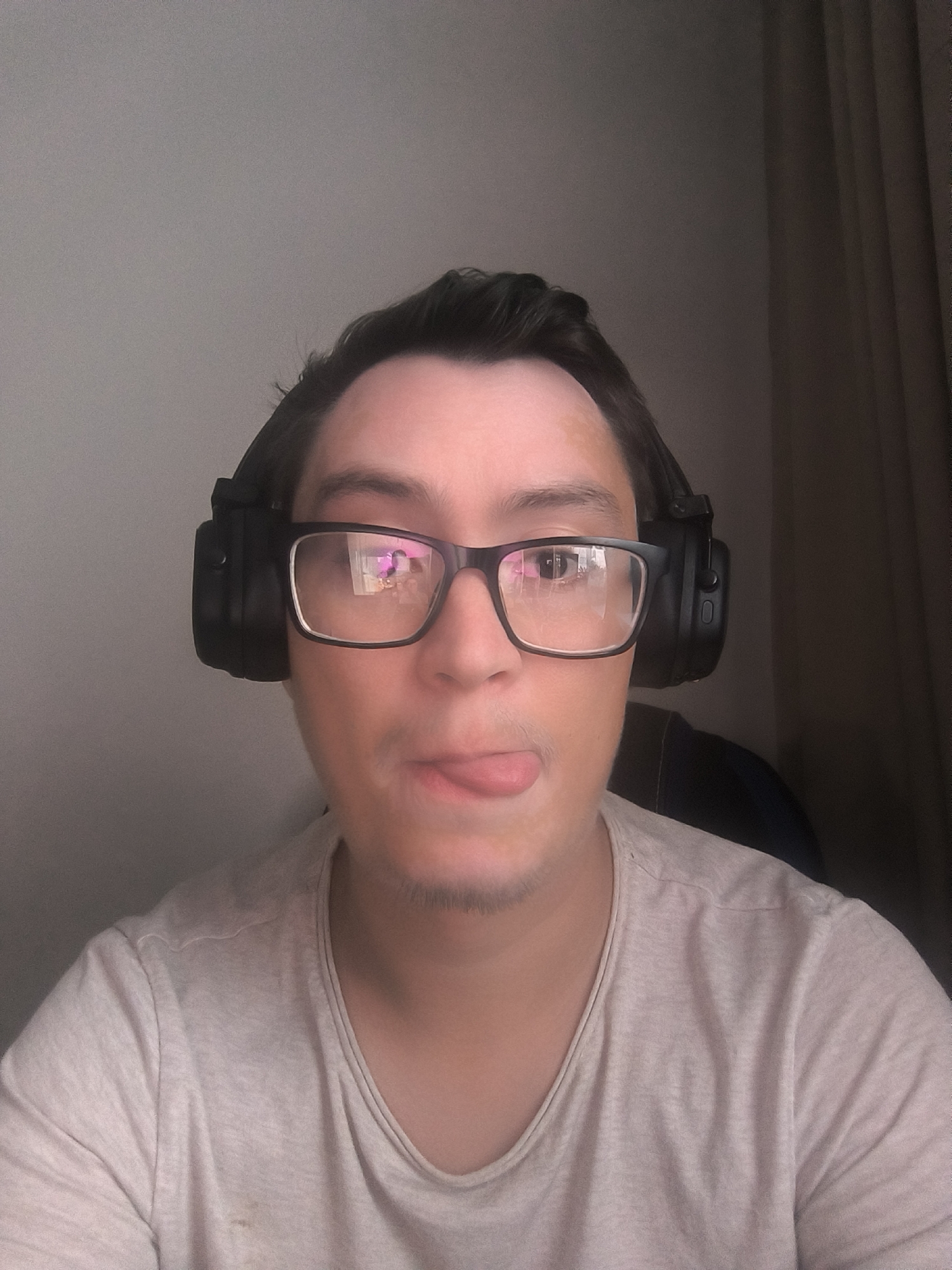 The image shows a close-up, head-and-shoulders shot of a person wearing large black headphones and glasses. The person has dark hair and is sticking their tongue out. They are wearing a light-colored t-shirt with some faint stains on it. The background is a plain, light-colored wall with a dark curtain on the right side. The lighting appears to be artificial and coming from the front. The scene is not in Kovrov, Russia.