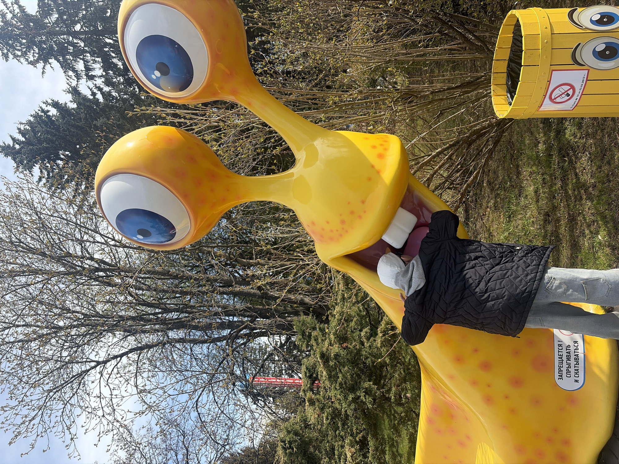 A child is standing in front of a large, yellow, slug-shaped sculpture with large blue eyes and big white teeth. The slug is located in an outdoor setting, likely a park or recreational area. A yellow trash receptacle with googly eyes is to the right. Behind the slug and the receptacle are trees and shrubs. The text on the sculpture is in Russian. The child is wearing a black quilted jacket, grey jeans and a white cap. The location is in Saint Petersburg, Russia.
